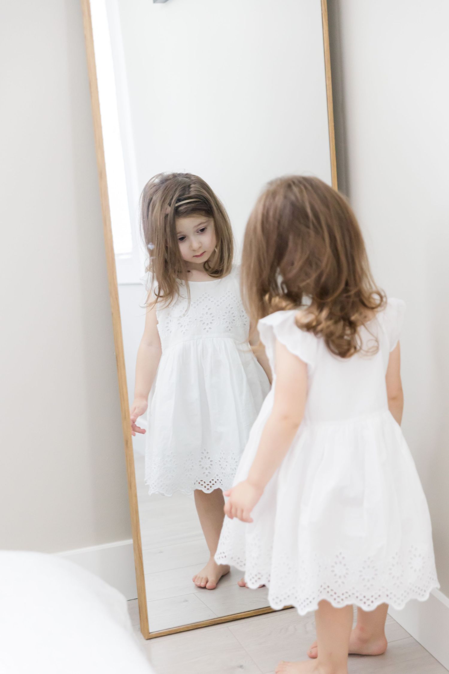 Child in white eyelet dress plays and looks at reflection in standing mirror with dog.
