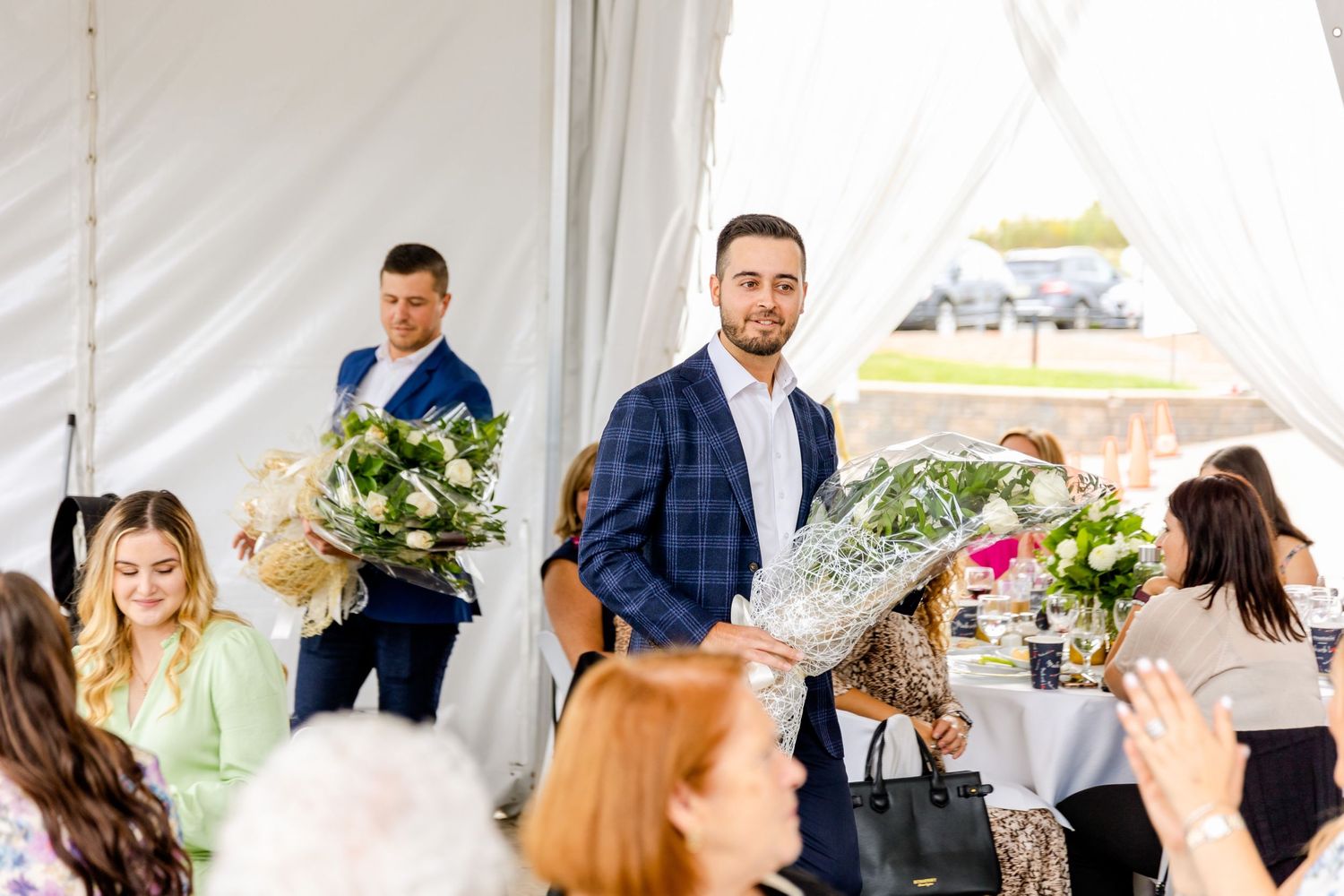 Guests celebrate at an outdoor event as attendees present floral bouquets.