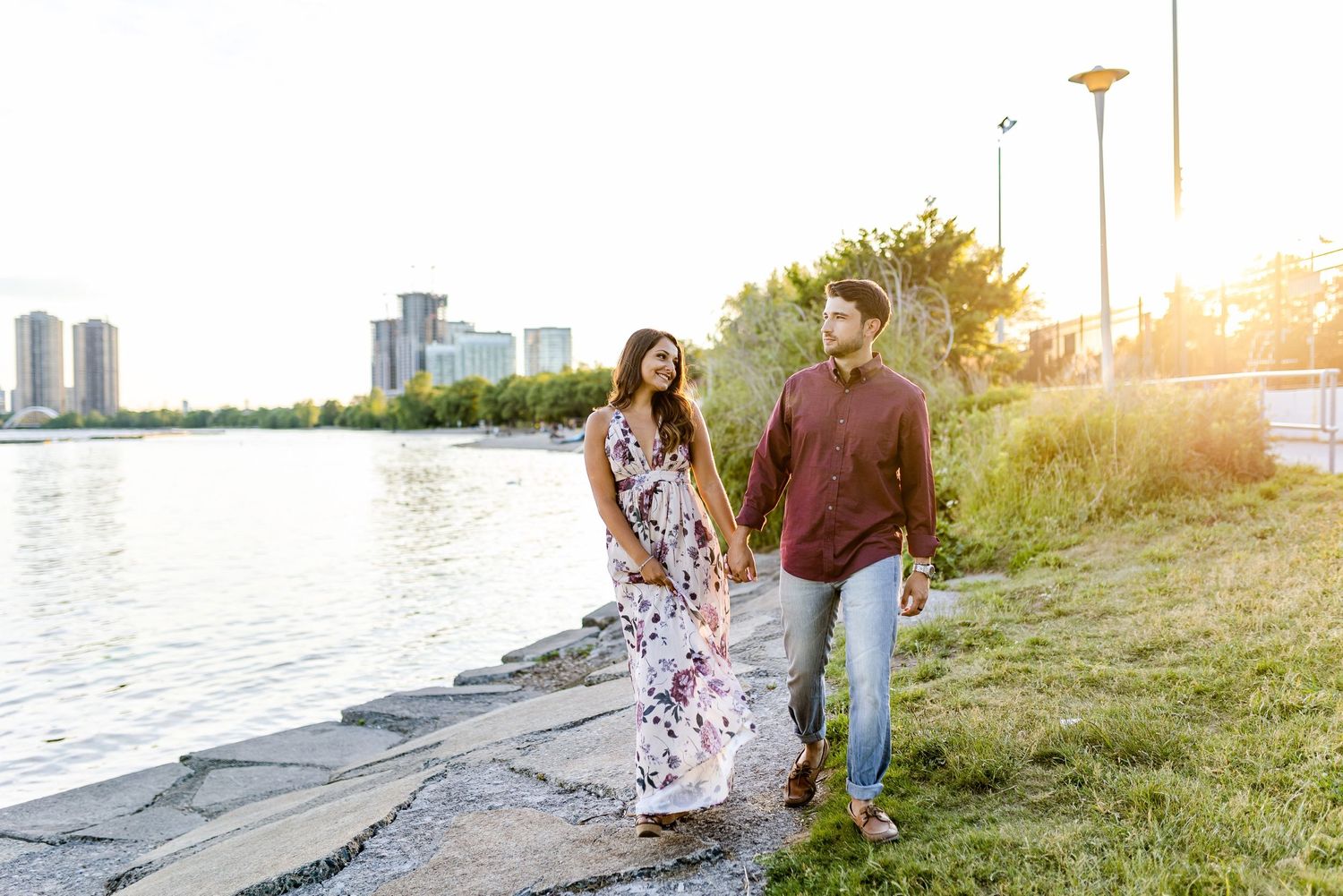 Couple walking along waterfront path at sunset wearing burgundy and floral outfits.