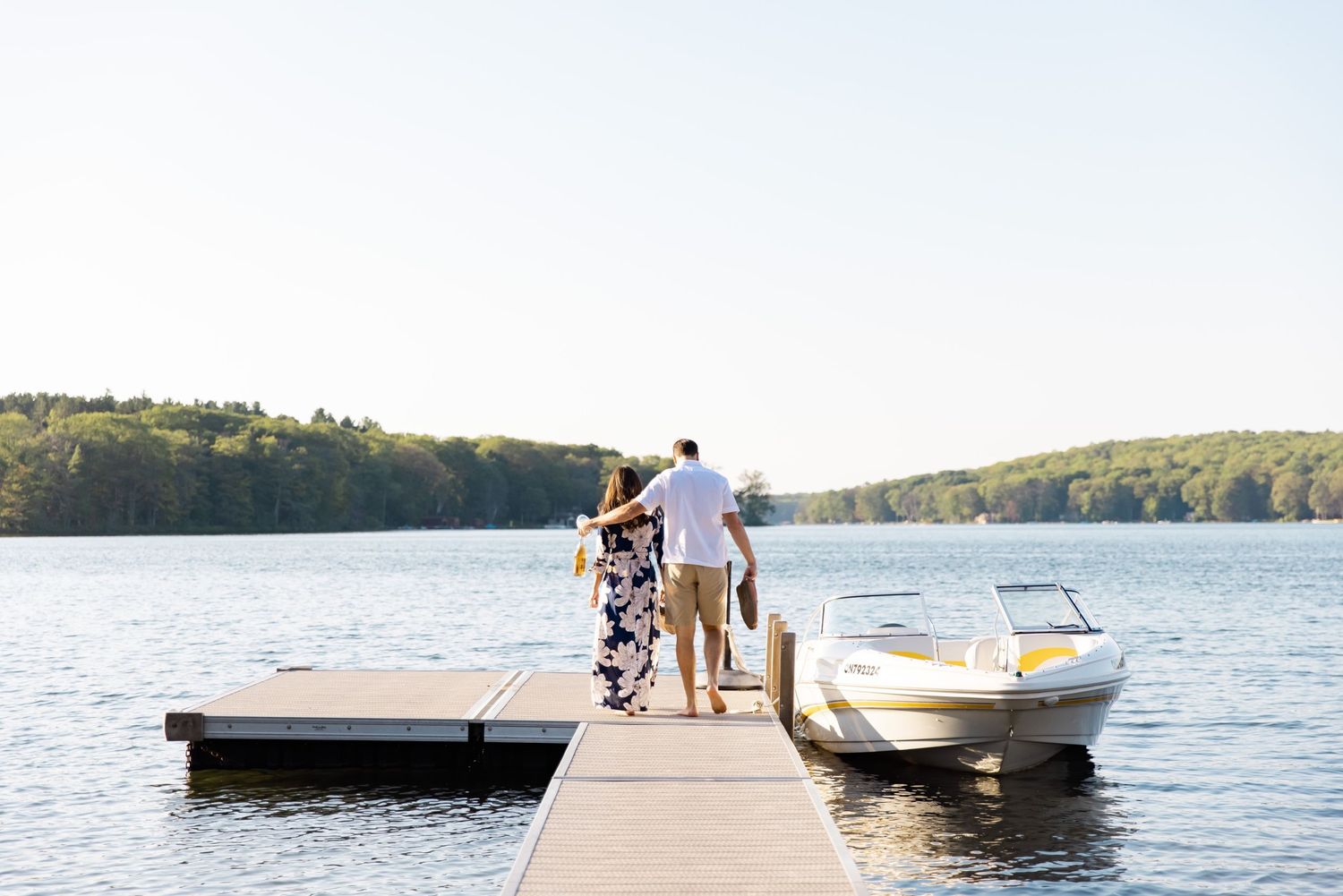 Couple strolling on lakeside dock with motorboat and wooded shoreline.