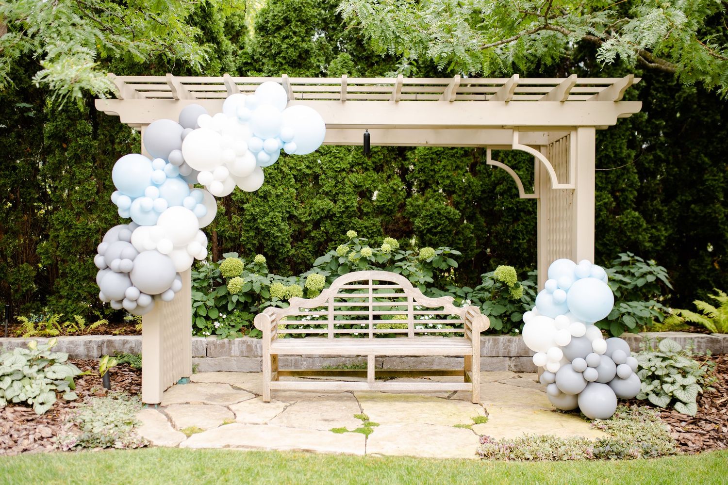 White and gray balloon arch decorates an elegant garden pergola with a classic wooden bench underneath.