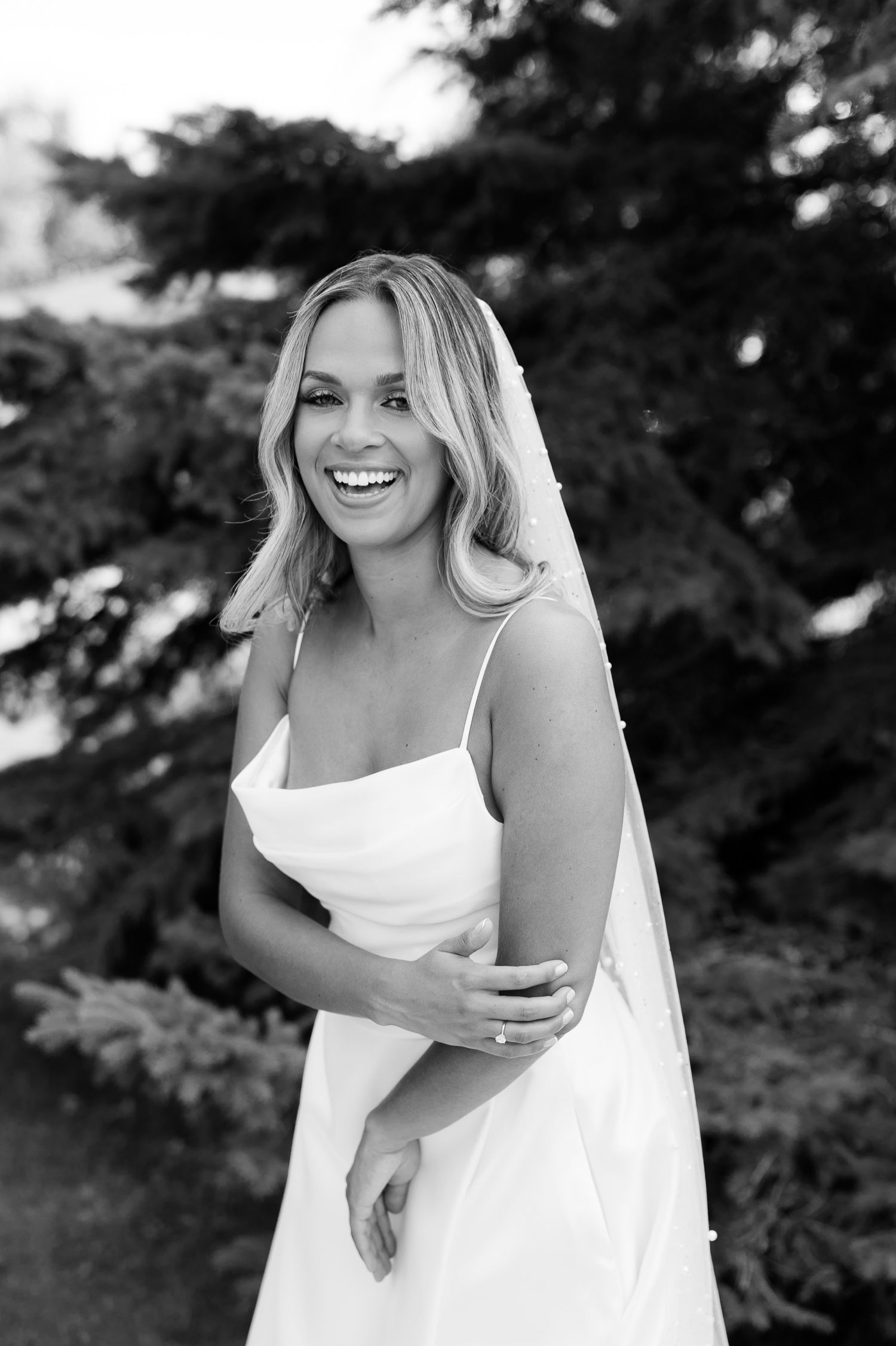 Black and white bridal portrait showing radiant smile against evergreen background.