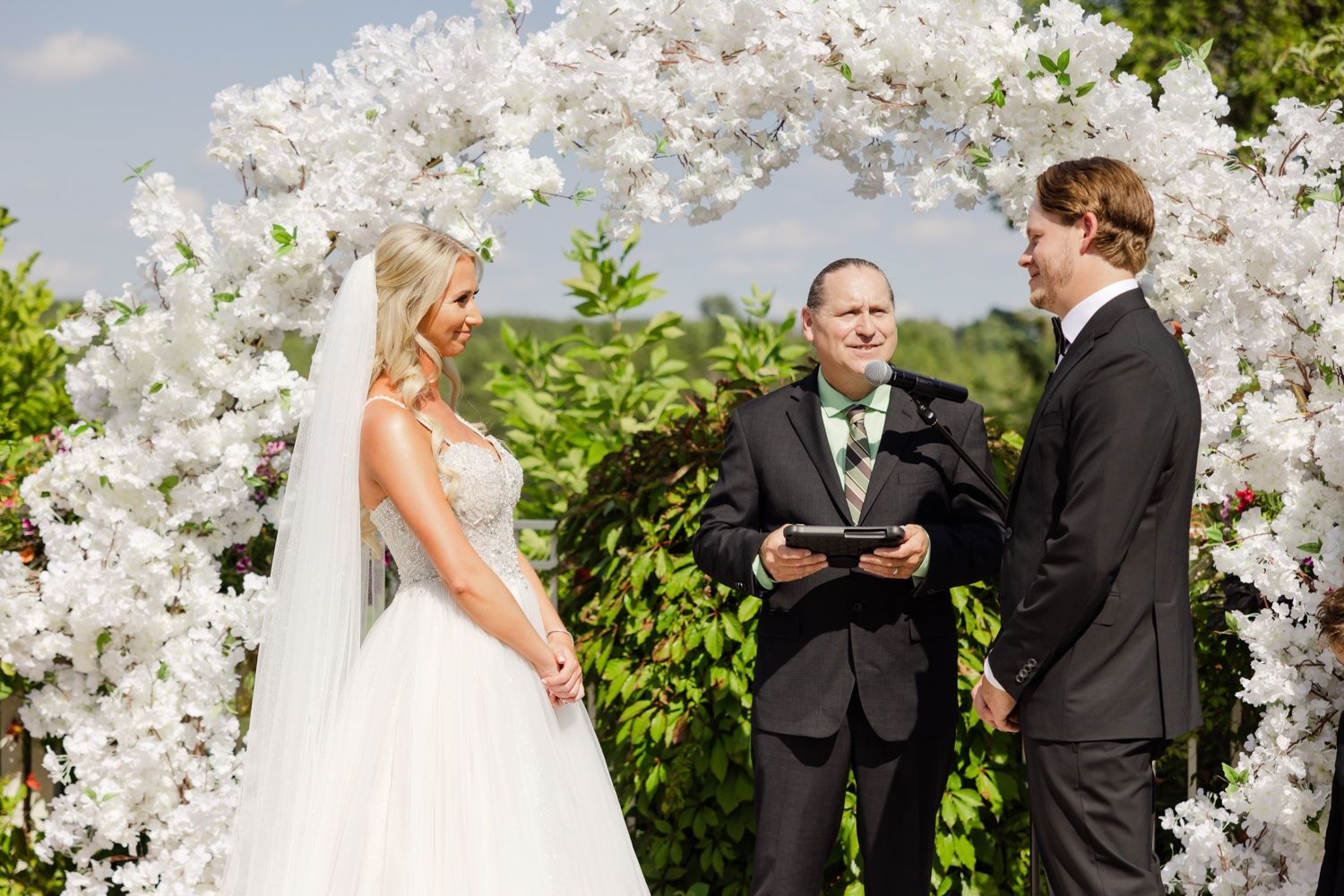 A wedding ceremony under a stunning white floral arch against a bright blue sky.