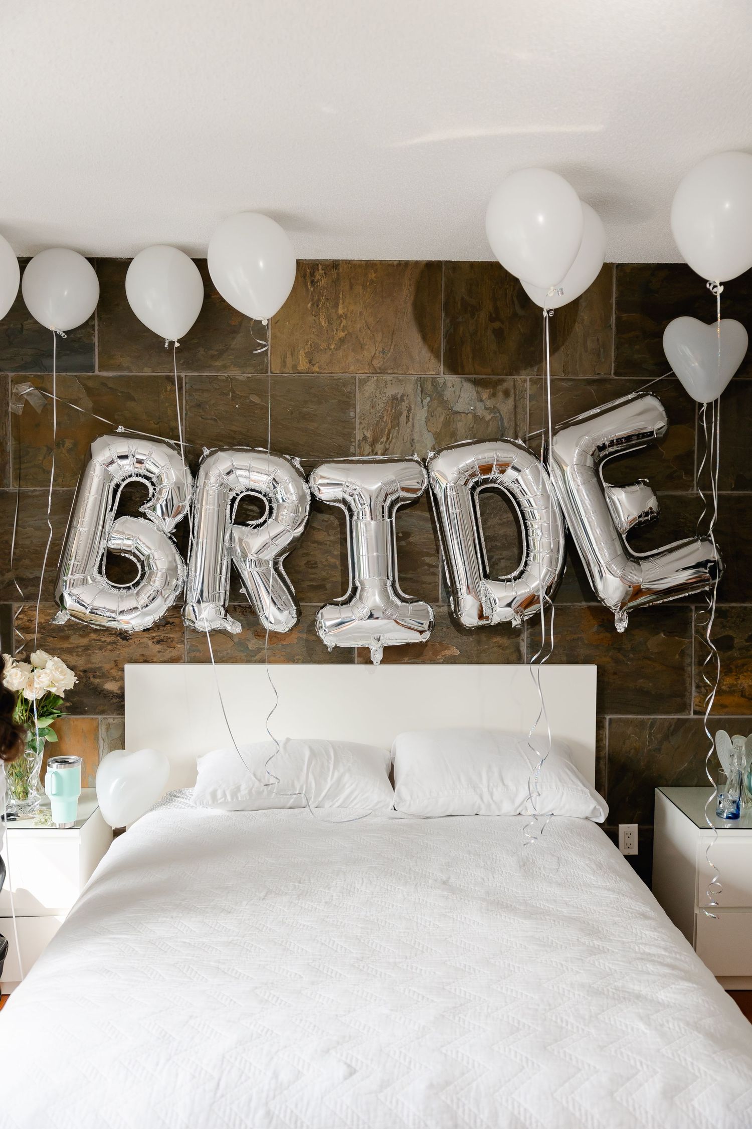 Silver balloon letters spelling BRIDE with white balloons above white bed against stone wall.