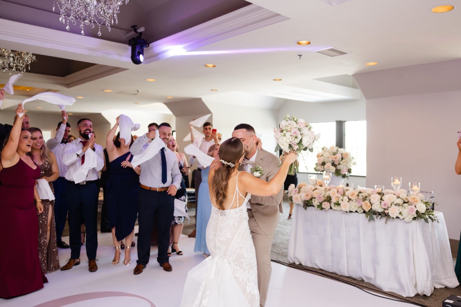 Wedding reception celebration with guests cheering and crystal chandeliers overhead.