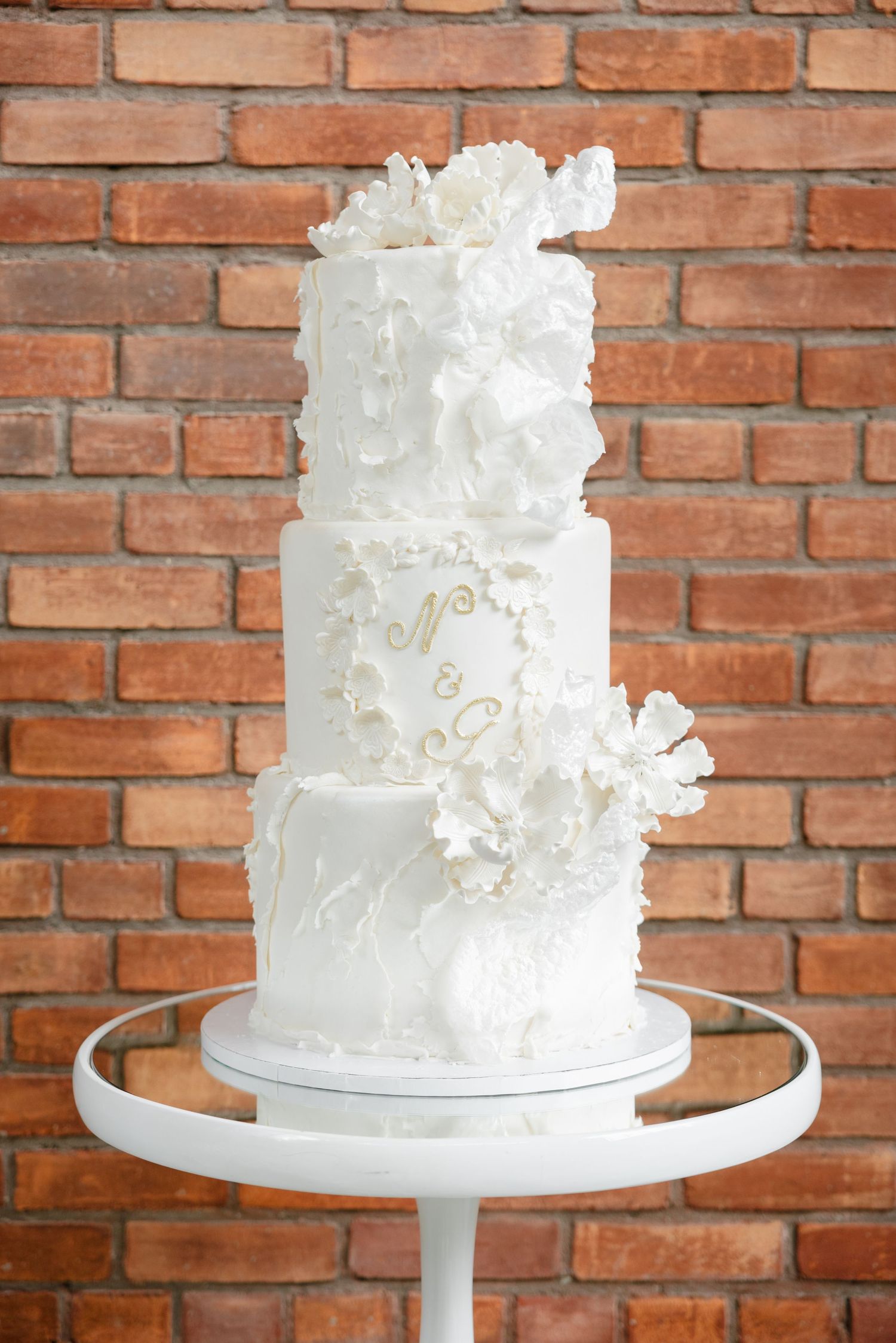 Three-tiered white wedding cake with delicate sugar flowers against brick wall.