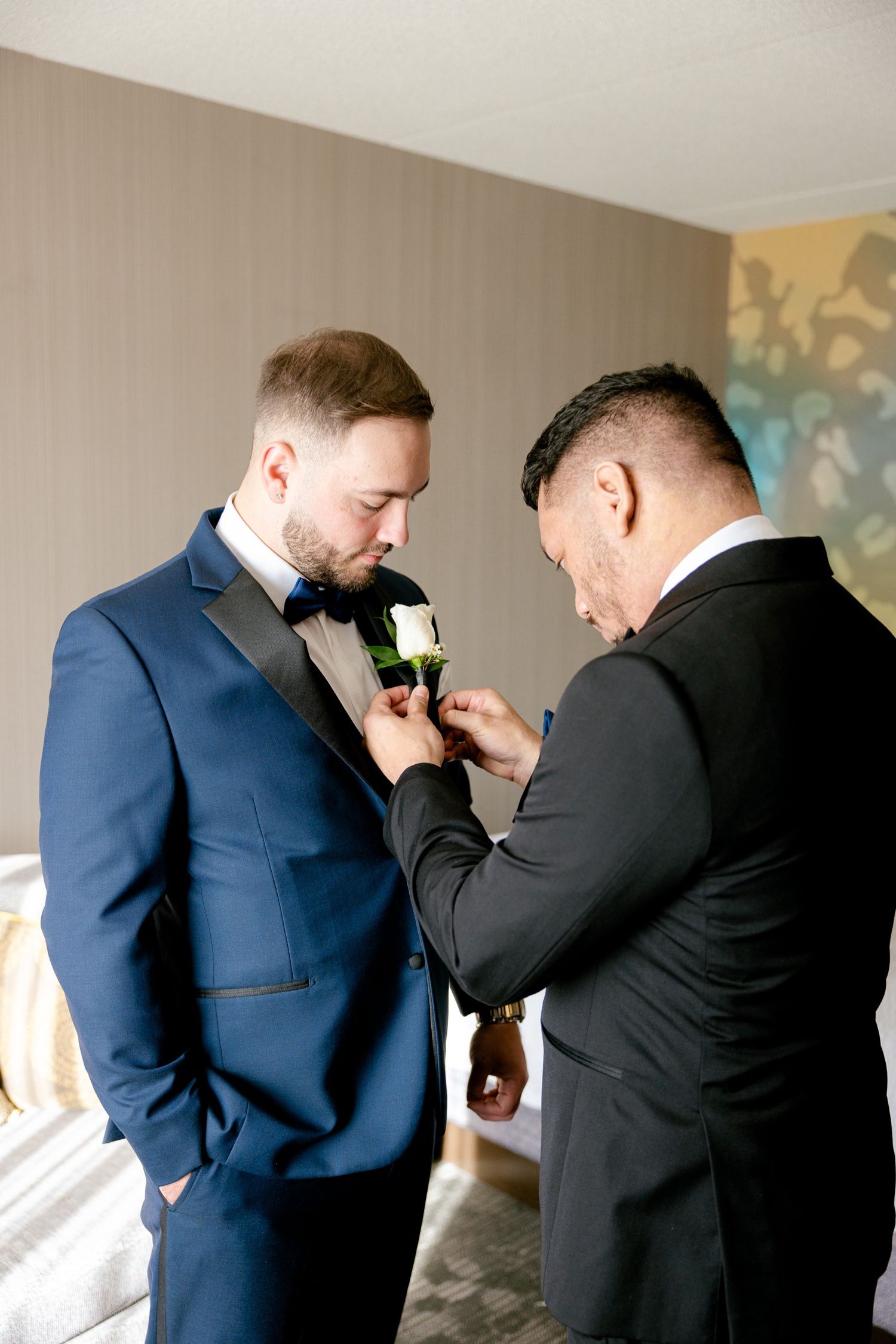 A groomsman helps adjust a white boutonniere on the groom's navy blue suit jacket before the wedding ceremony.