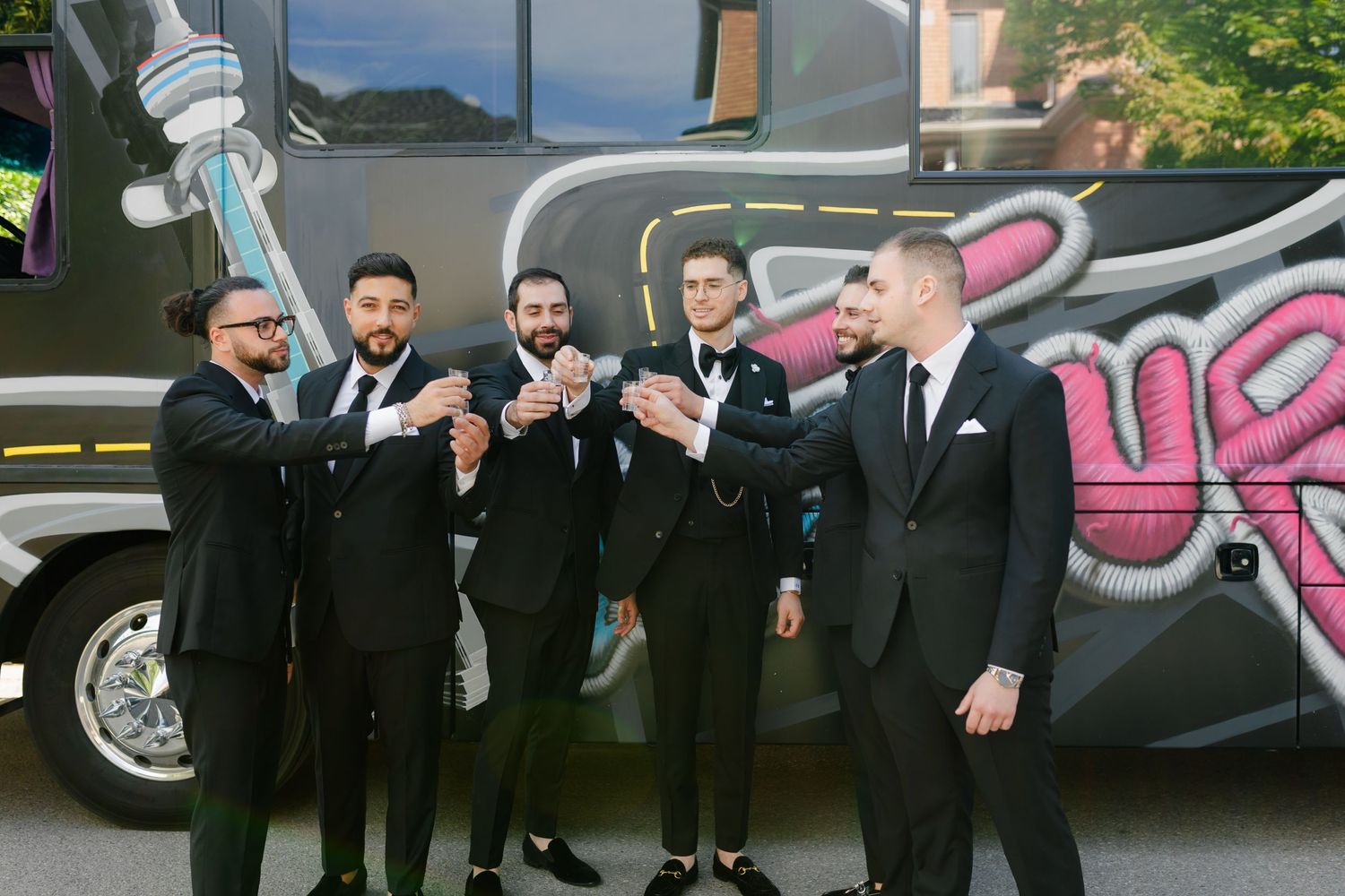 Groomsmen in black suits pose together in front of a party bus with graffiti art.