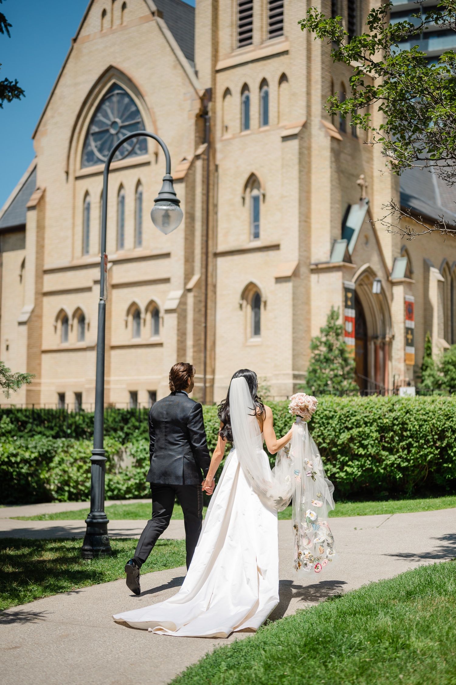 A couple walks toward a Gothic-style church with cream colored stone and arched windows on their wedding day.