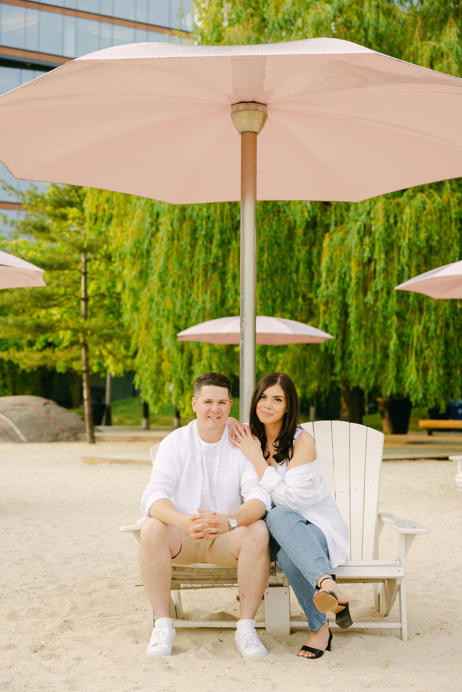 Couple poses together on beach chair under pink umbrella with lush greenery backdrop.