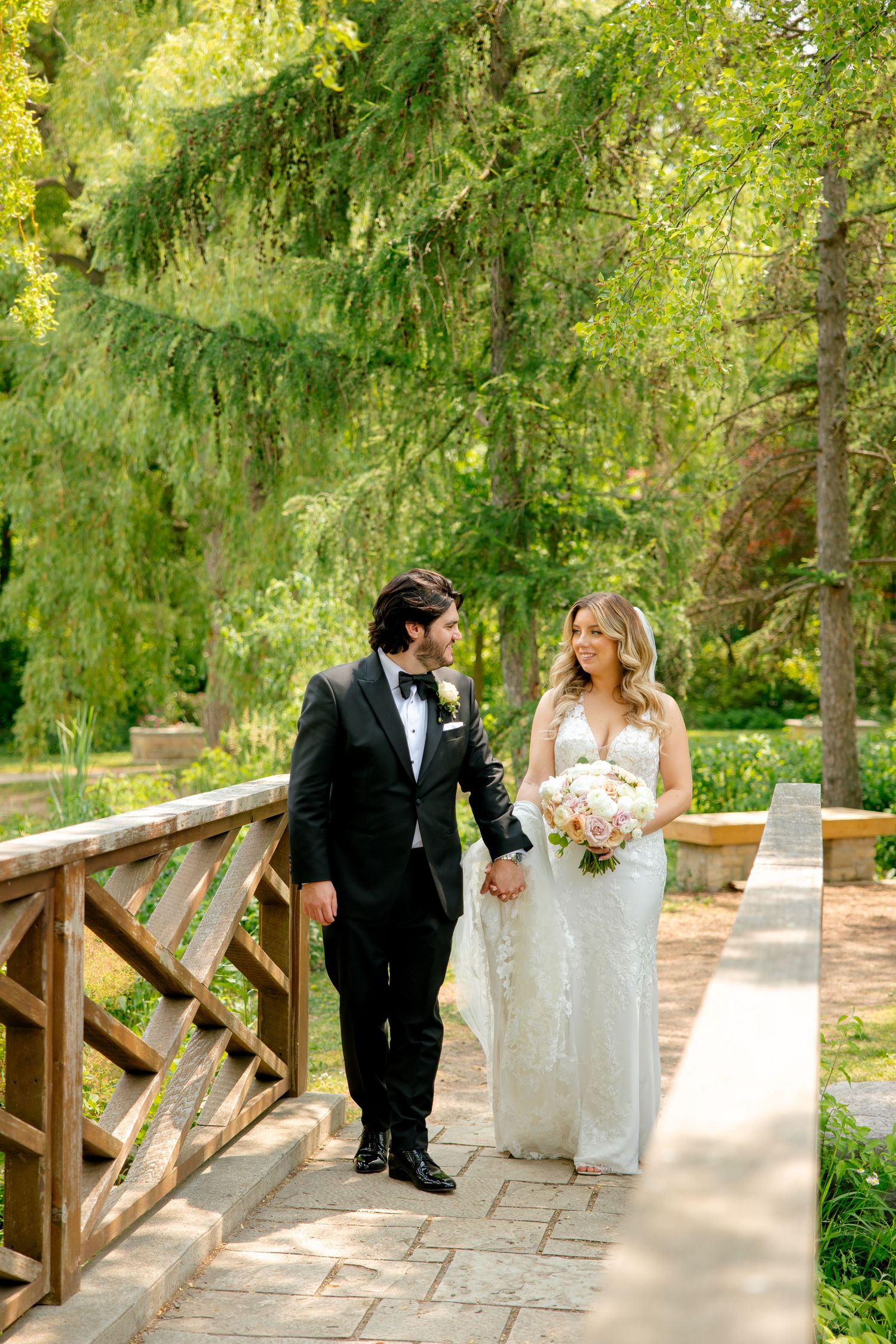 Bride and groom walk hand in hand across rustic bridge surrounded by spring greenery.