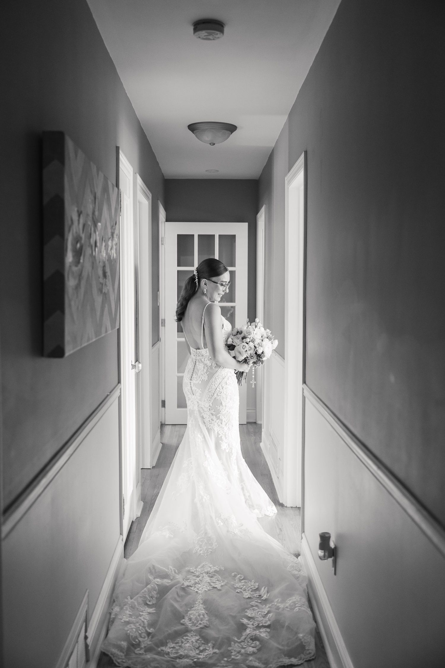 Black and white photo of a bride in a detailed lace gown standing in a hallway.