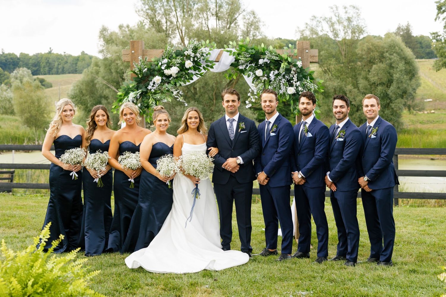 Wedding party gathered under a white floral arch against a scenic countryside backdrop.