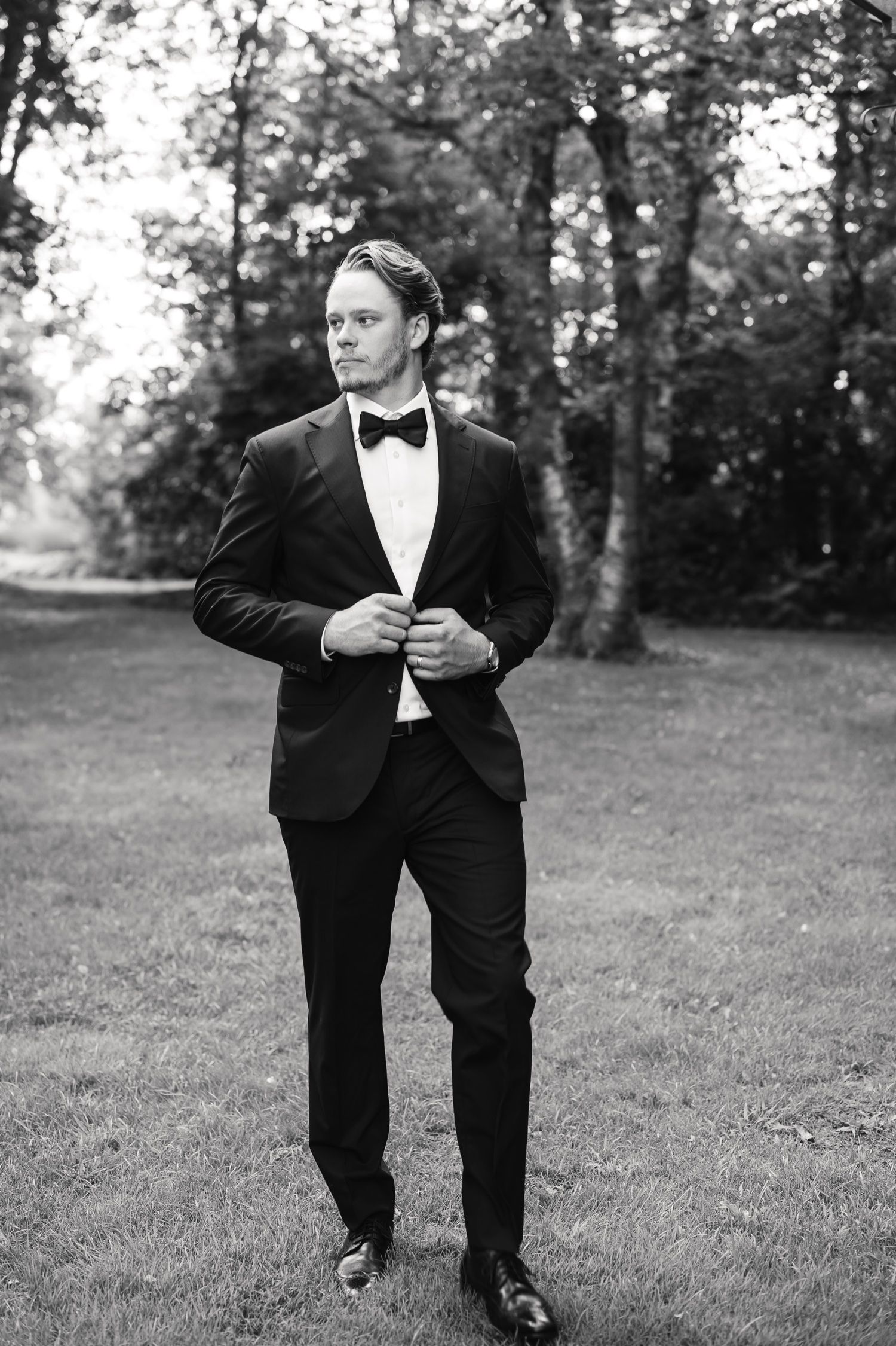 Black and white portrait of a groom adjusting his suit jacket in an outdoor garden setting.