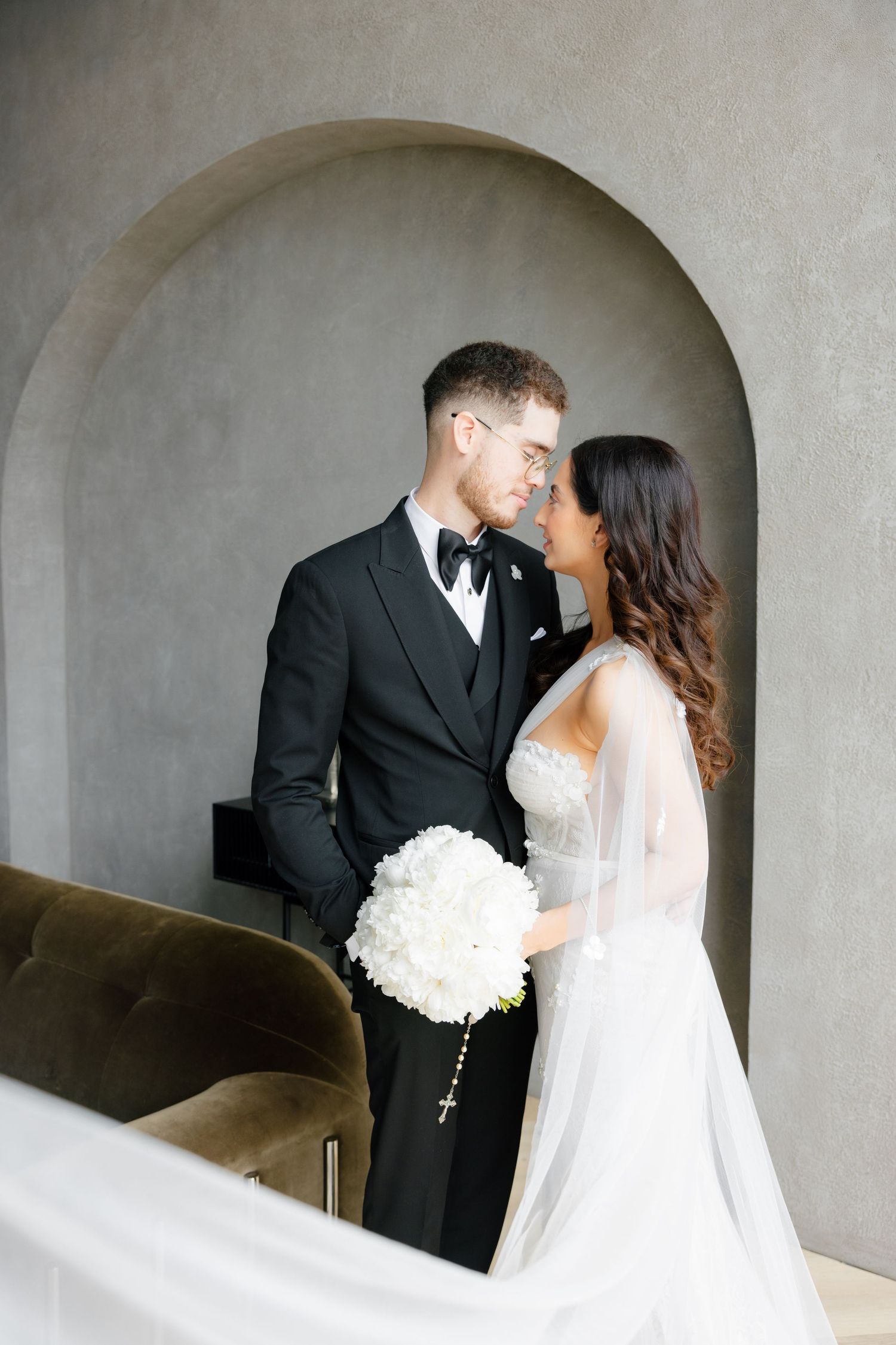 Bride in white dress holds bouquet while sharing intimate moment with groom under archway.