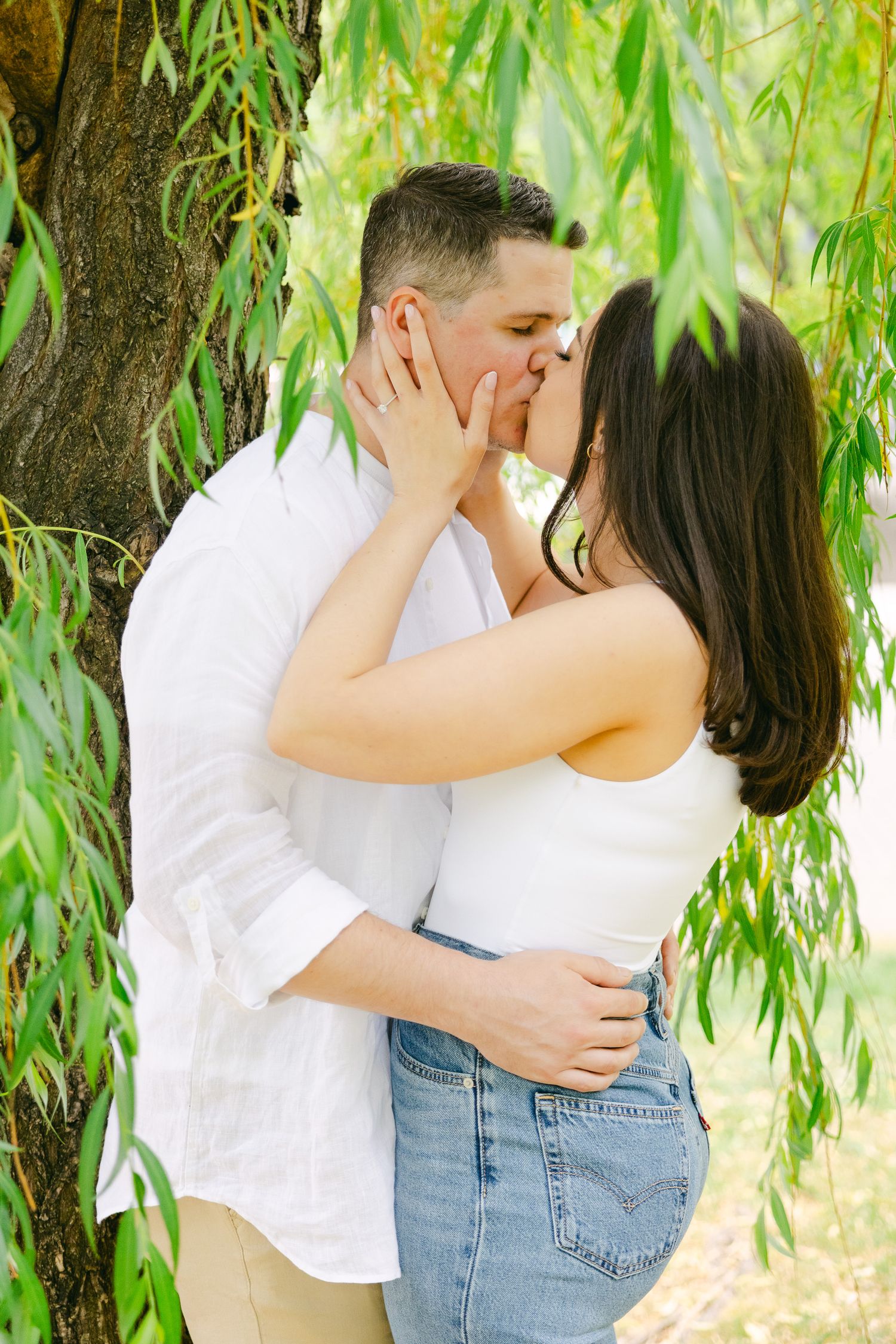 A romantic moment shared beneath hanging willow branches on a sunny day.