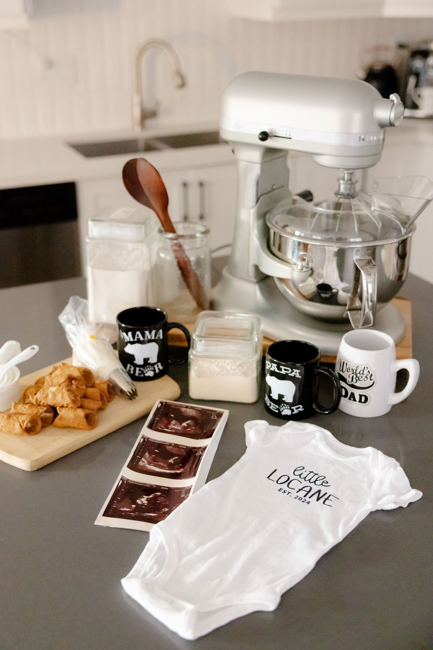 Kitchen countertop with baking items, mugs, and baby onesie suggesting pregnancy announcement.