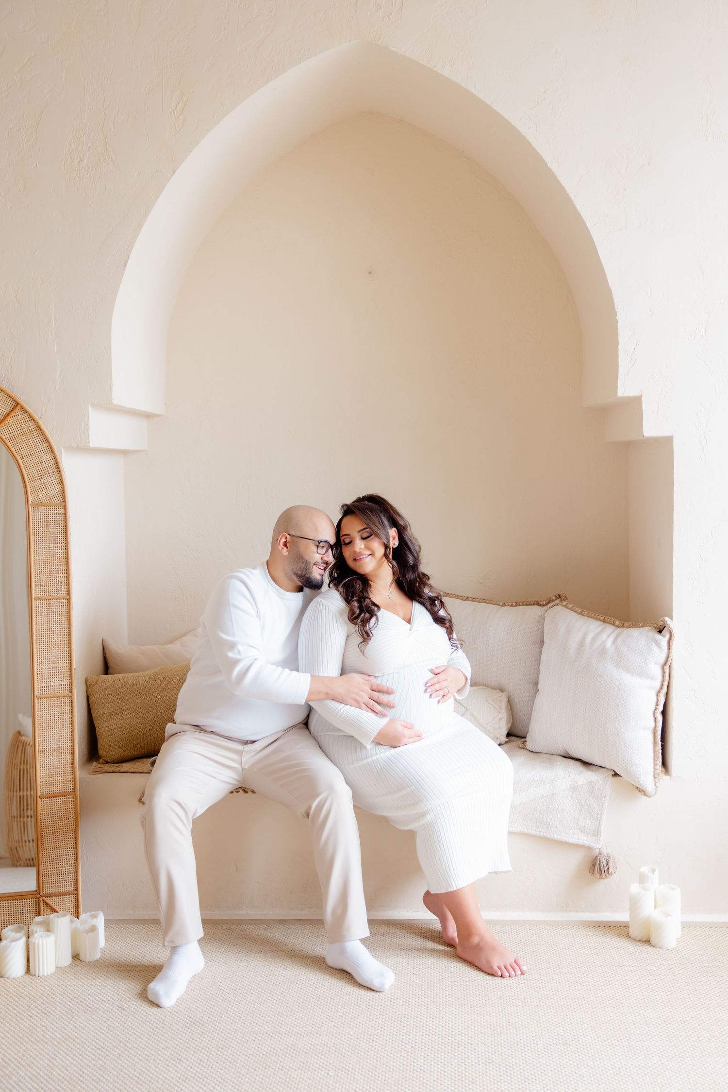 Couple dressed in white sitting together in an arched alcove with white cushions and romantic candlelight.