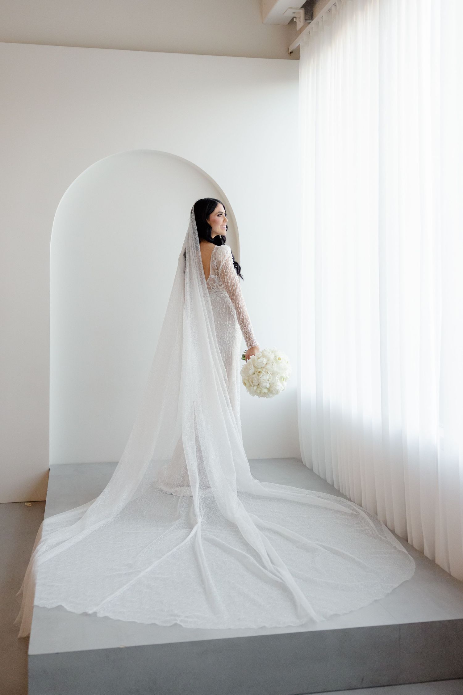 Bride in delicate lace gown stands in arched alcove with flowing cathedral veil.