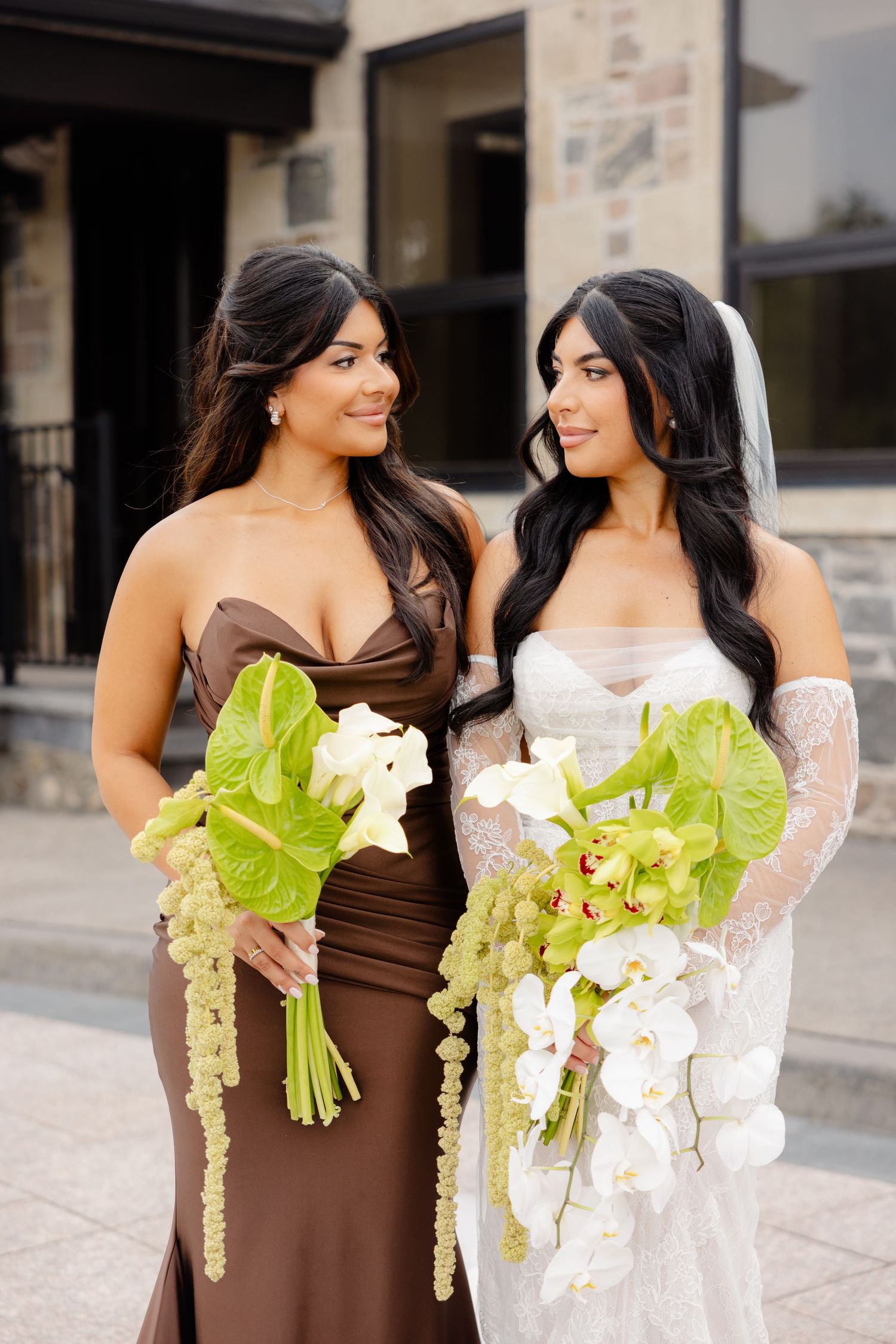 Two bridesmaids hold elegant floral bouquets featuring white orchids and green accents in brown and white dresses.