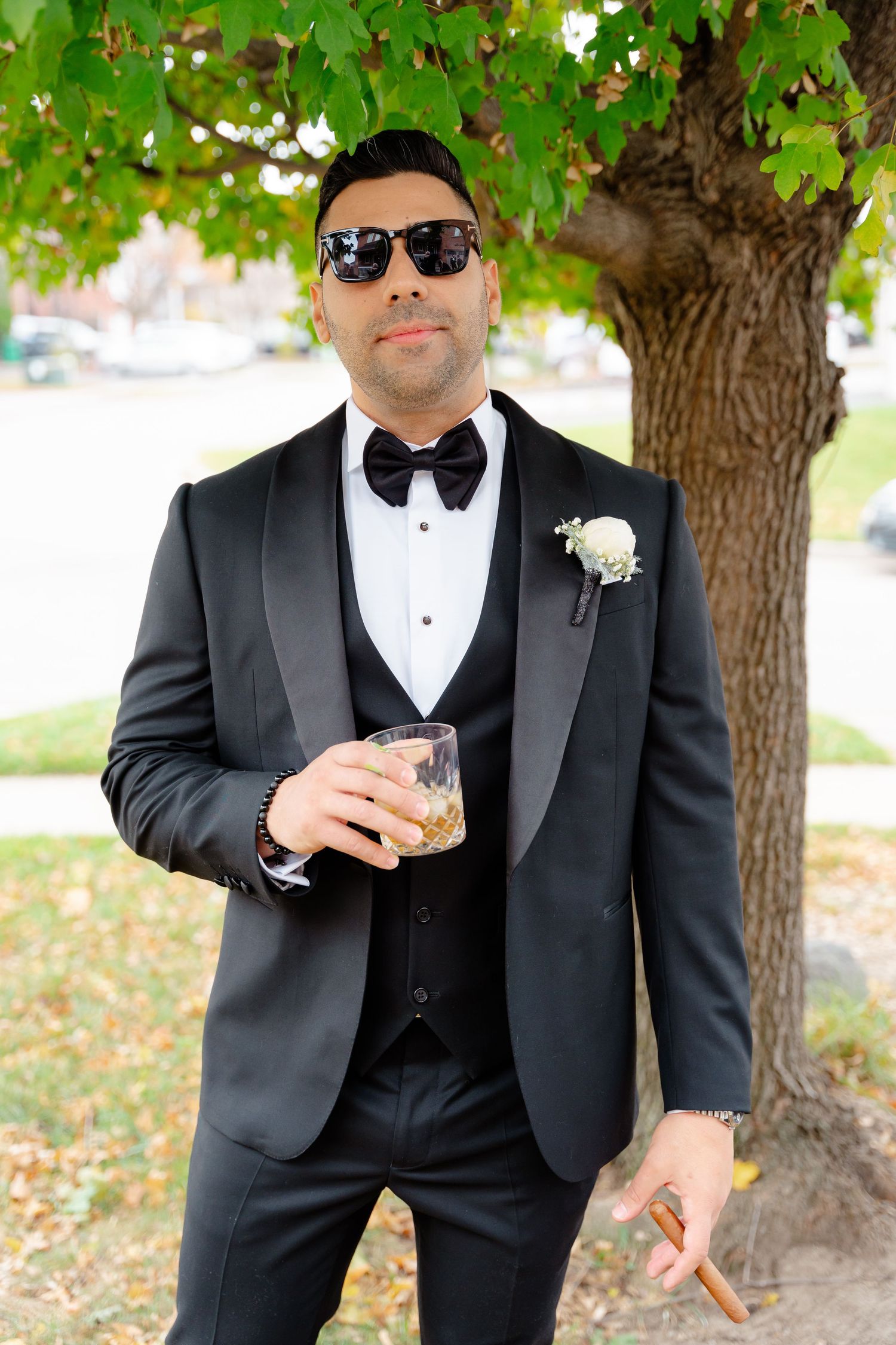 Stylish groomsman in black tuxedo posing with drink under autumn tree.
