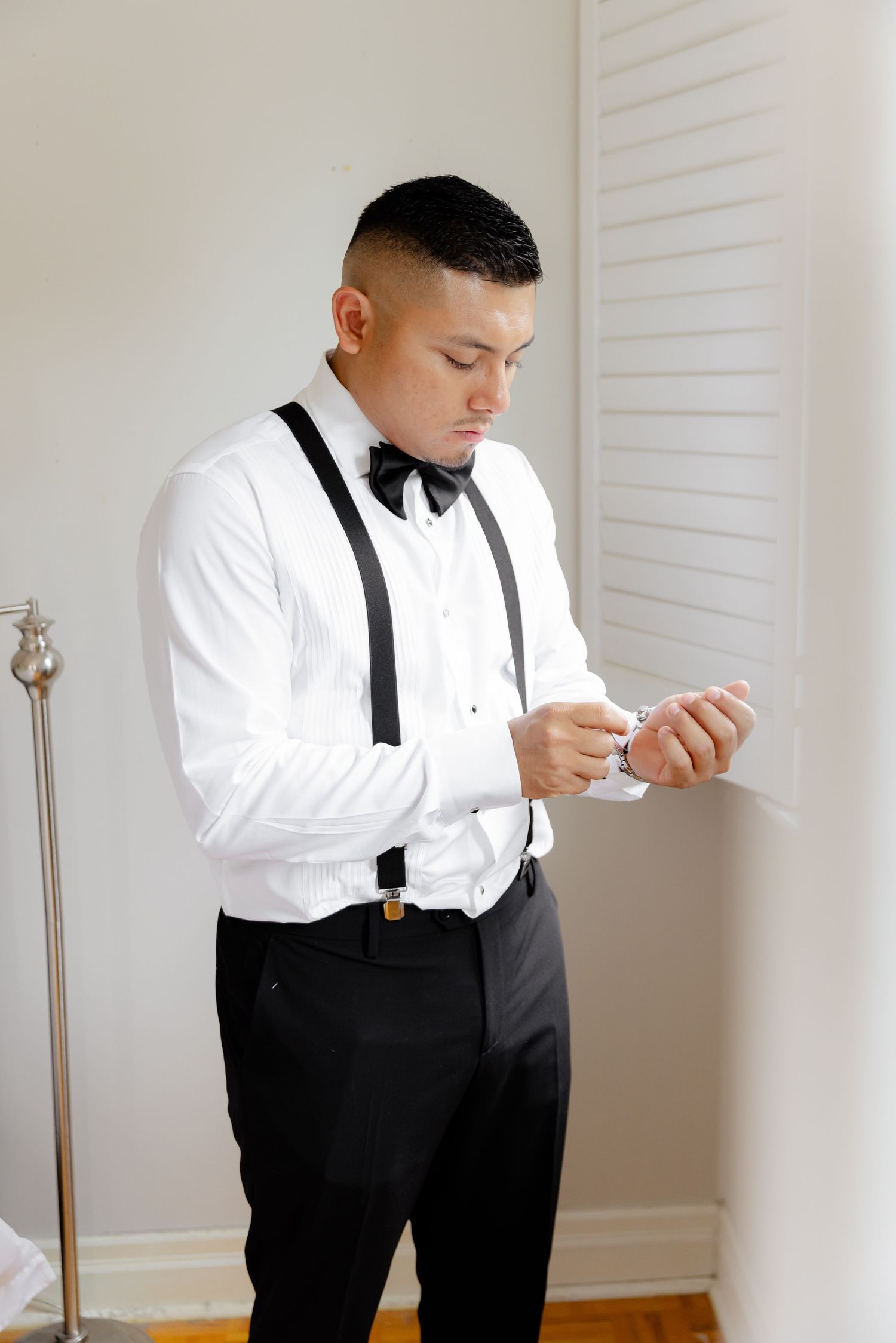 Person in formal attire with black suspenders and bow tie adjusts cuffs while getting ready in minimalist setting.