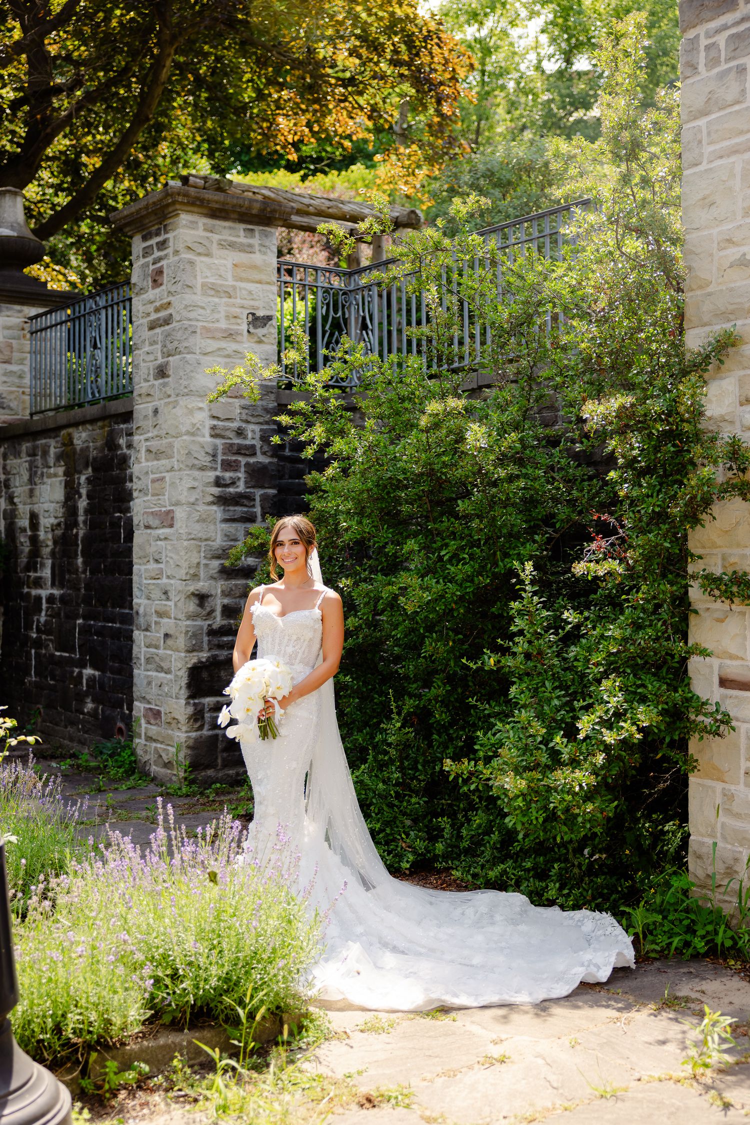 A white lace wedding dress with a long flowing train photographed in a garden setting with stone walls and lavender flowers.