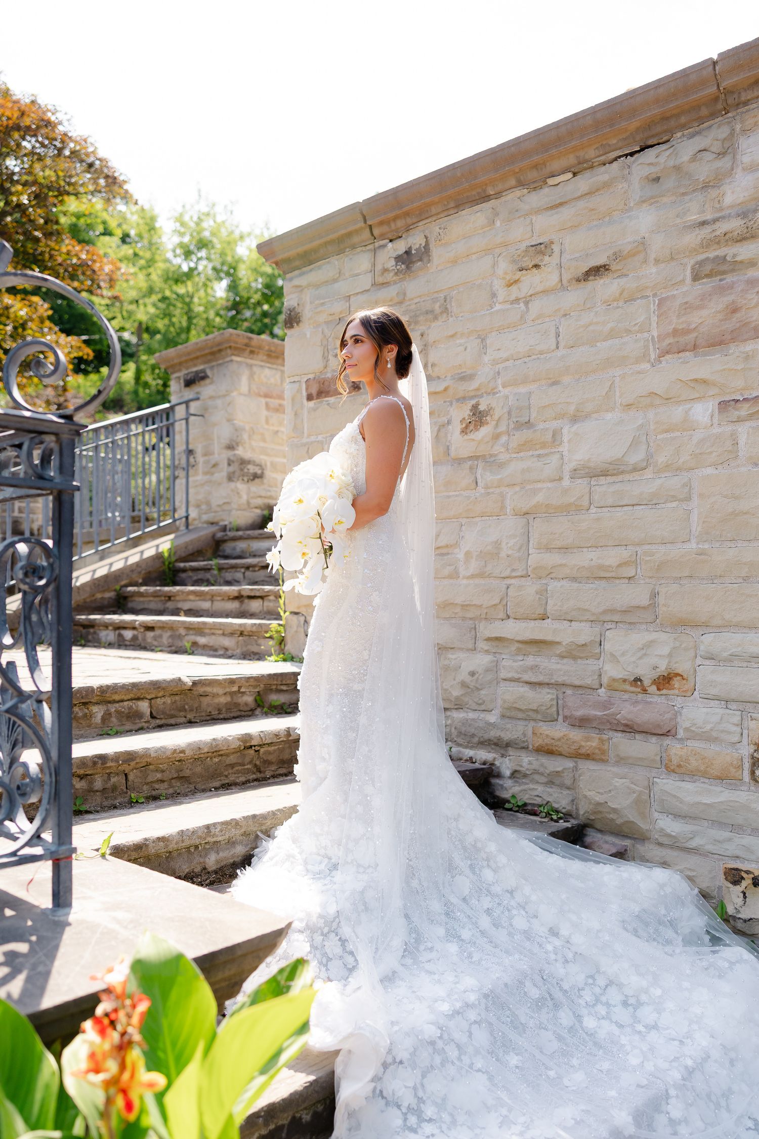 Elegant wedding dress with flowing train cascades down stone staircase in outdoor garden setting.