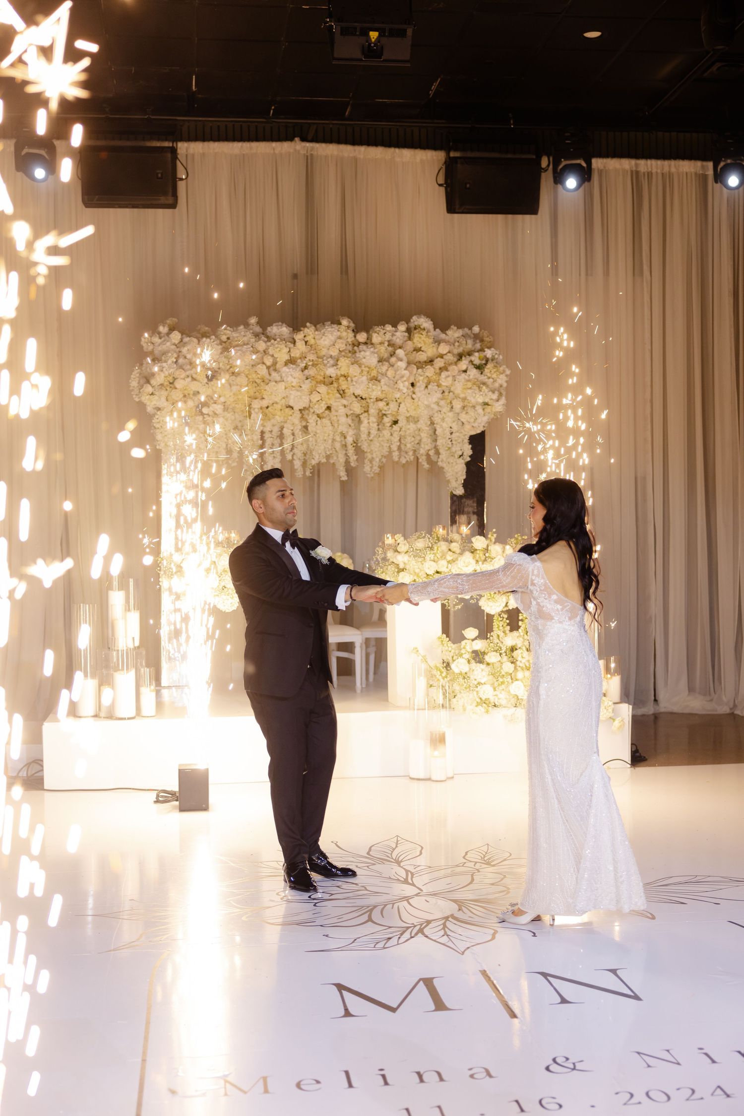 Dancing couple with sparkler effects under white floral arch on personalized dance floor.