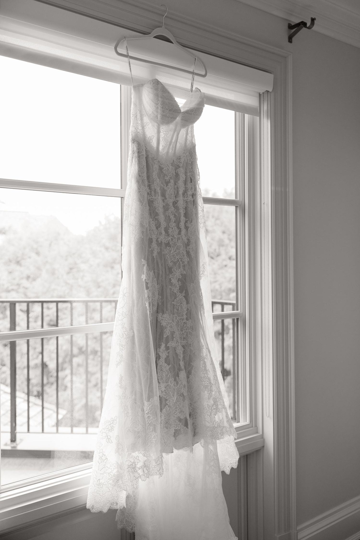 A delicate lace wedding dress hangs by a window in black and white, with natural light filtering through.