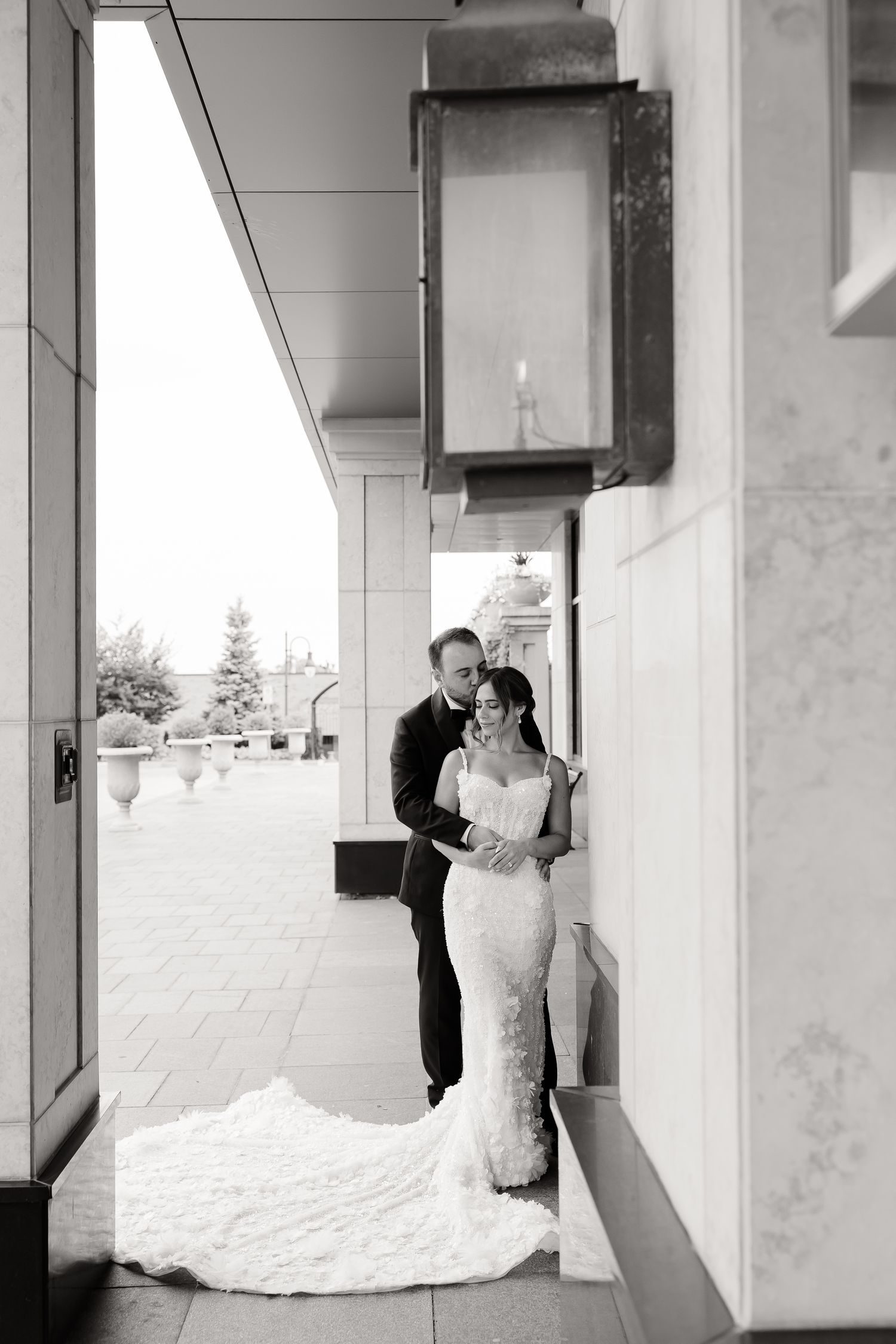 Black and white wedding portrait of couple standing in elegant outdoor corridor with vintage lantern lighting.