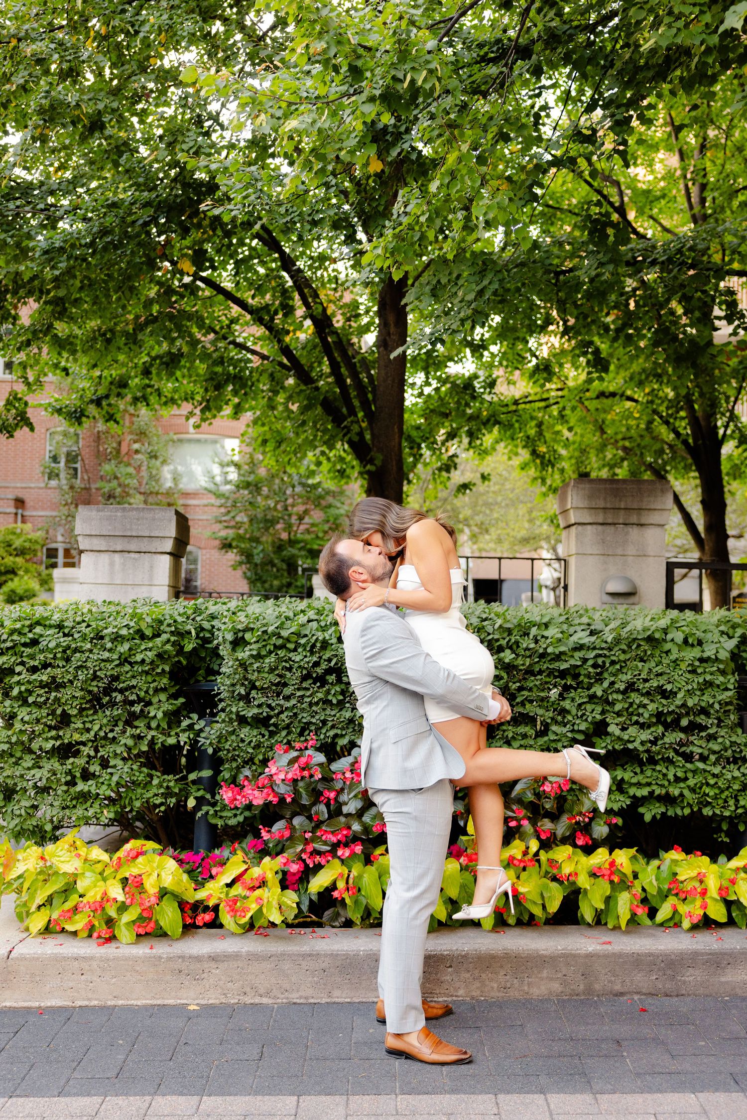 A joyful couple embraces in a lifting pose against a backdrop of trees and flowers.