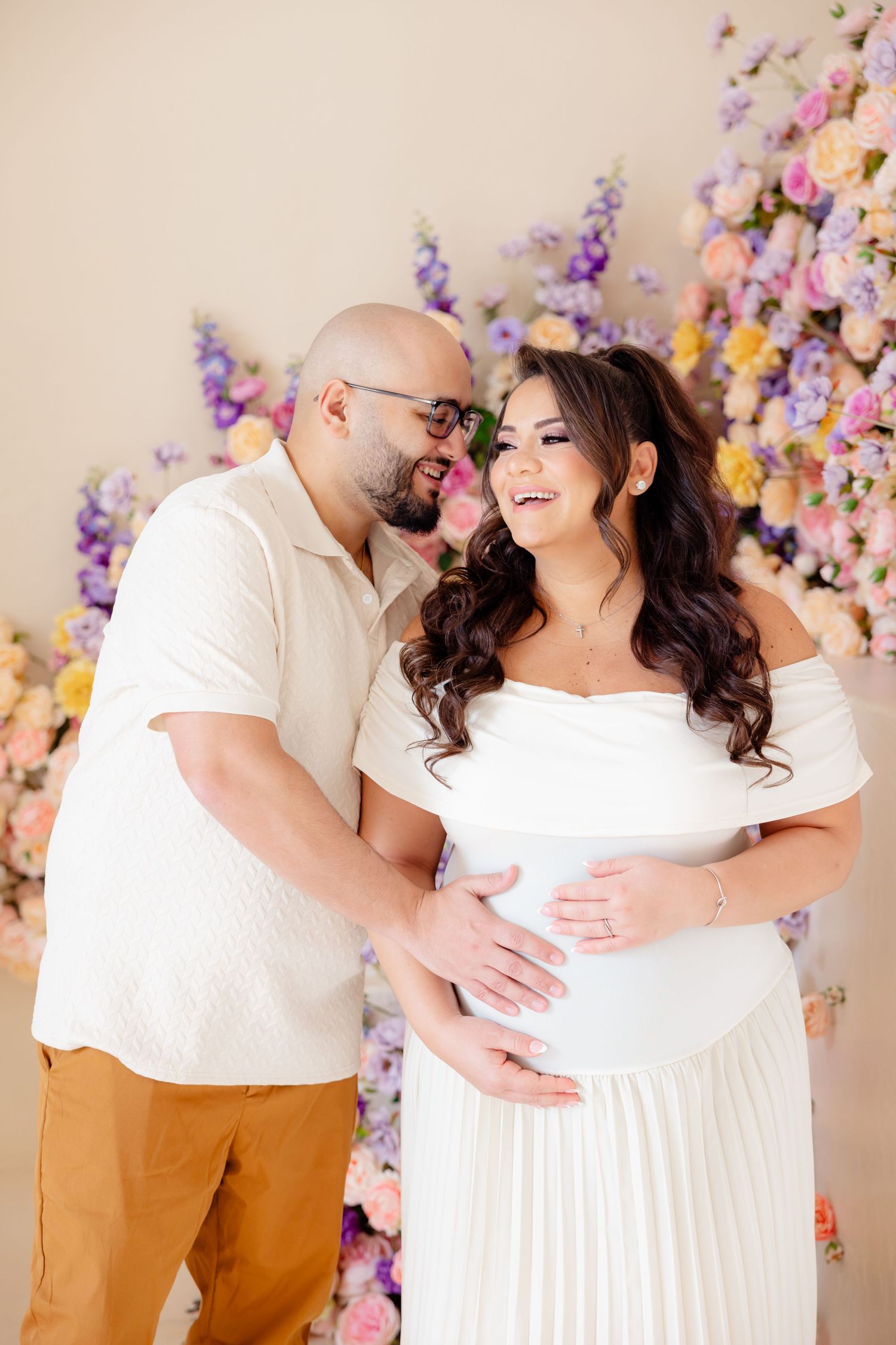 Expecting couple shares tender moment in front of floral wall backdrop during maternity photo shoot.