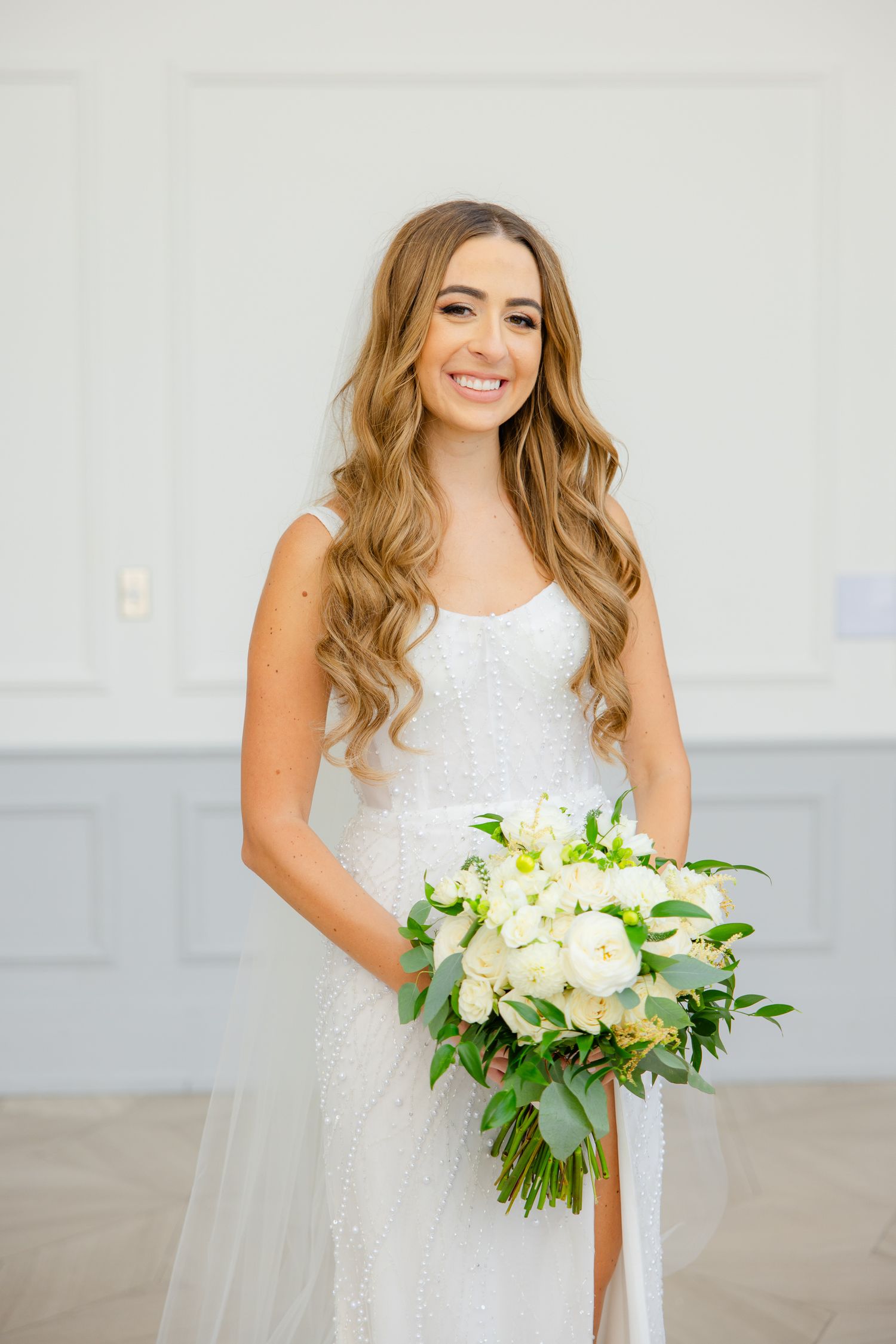 Radiant bride poses with white floral bouquet in elegant fitted wedding dress with flowing train.