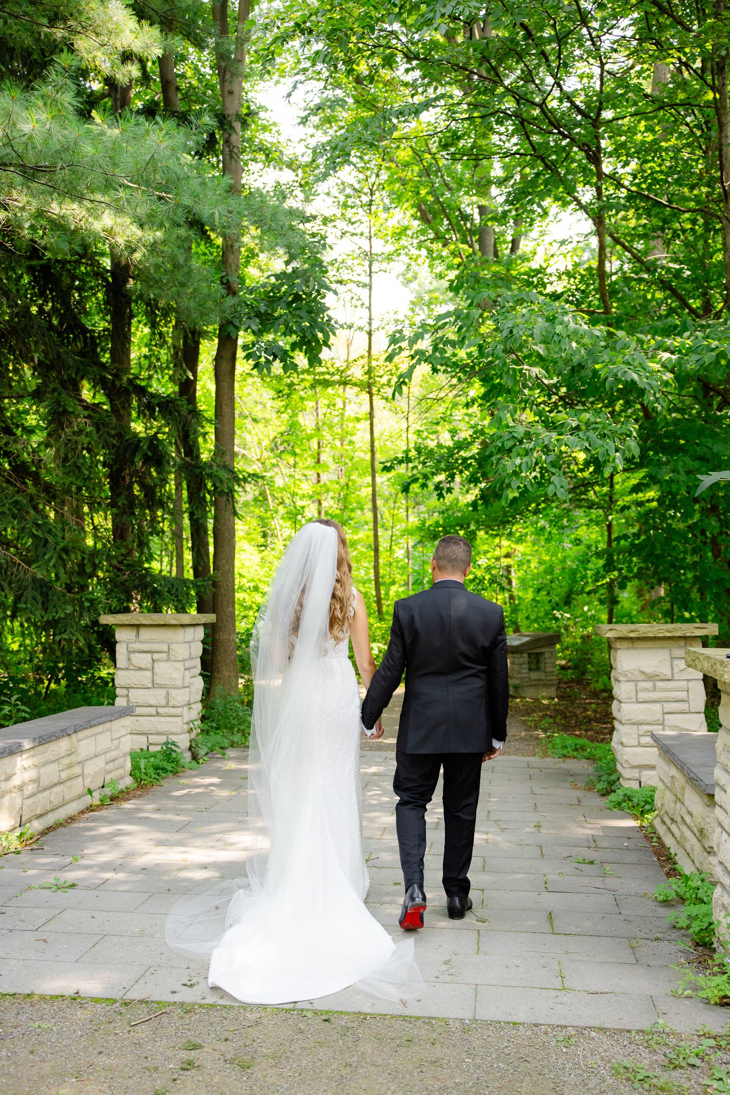Bride and groom walking away on stone pathway surrounded by green trees.