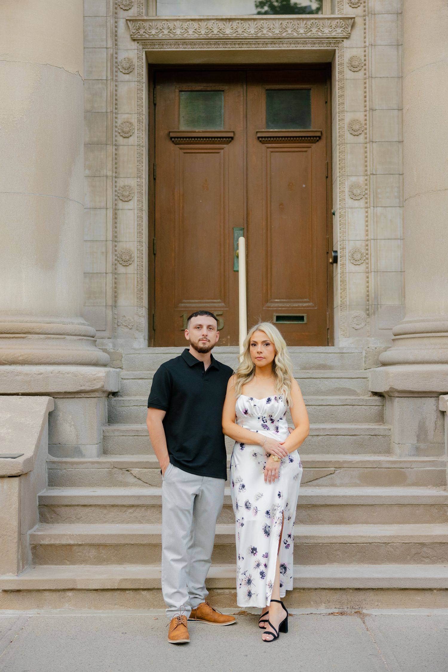 A couple stands together on stone steps in front of an ornate wooden door.