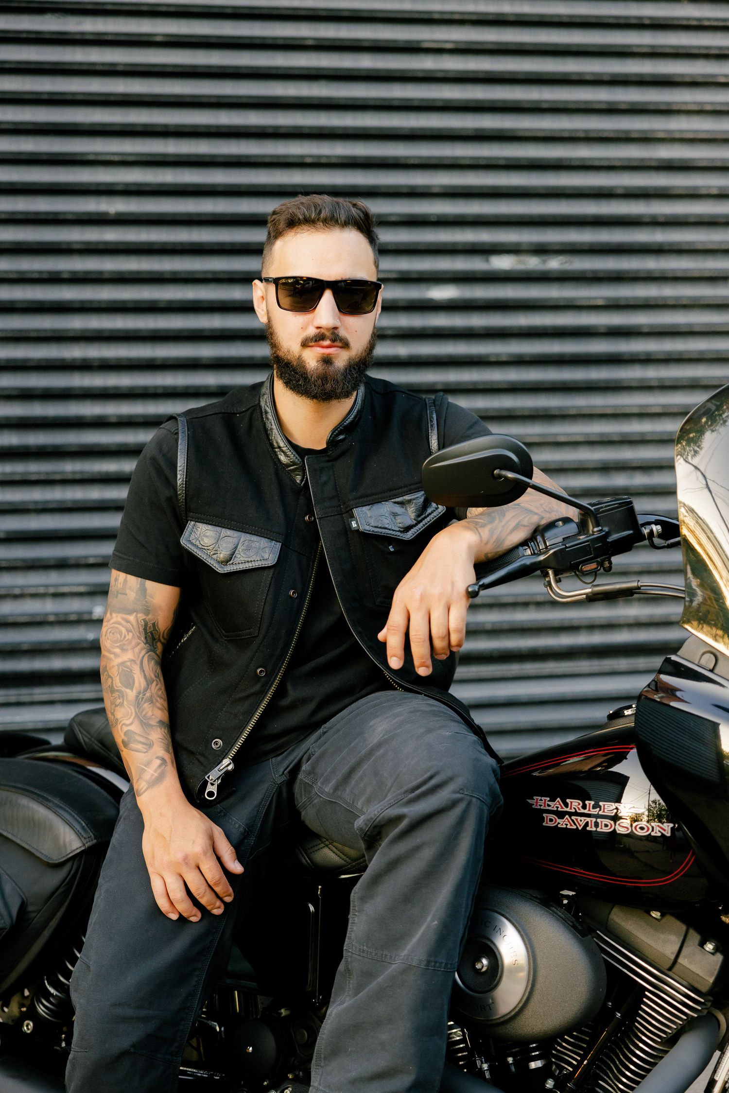 A motorcyclist in a black vest and sunglasses poses on their parked Harley Davidson against industrial siding.
