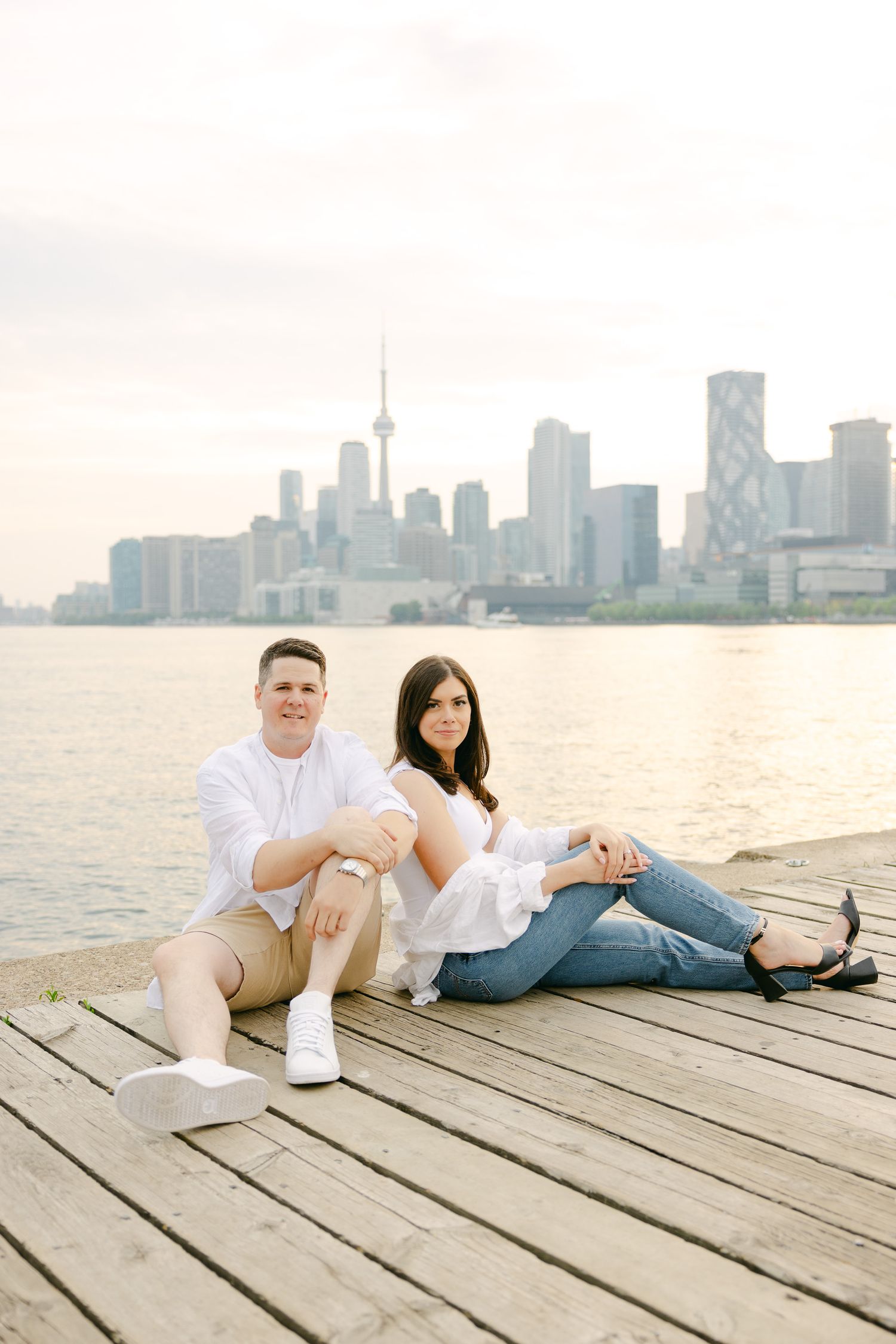 A casual engagement photo of a couple relaxing on a wooden dock with Toronto's skyline behind them.