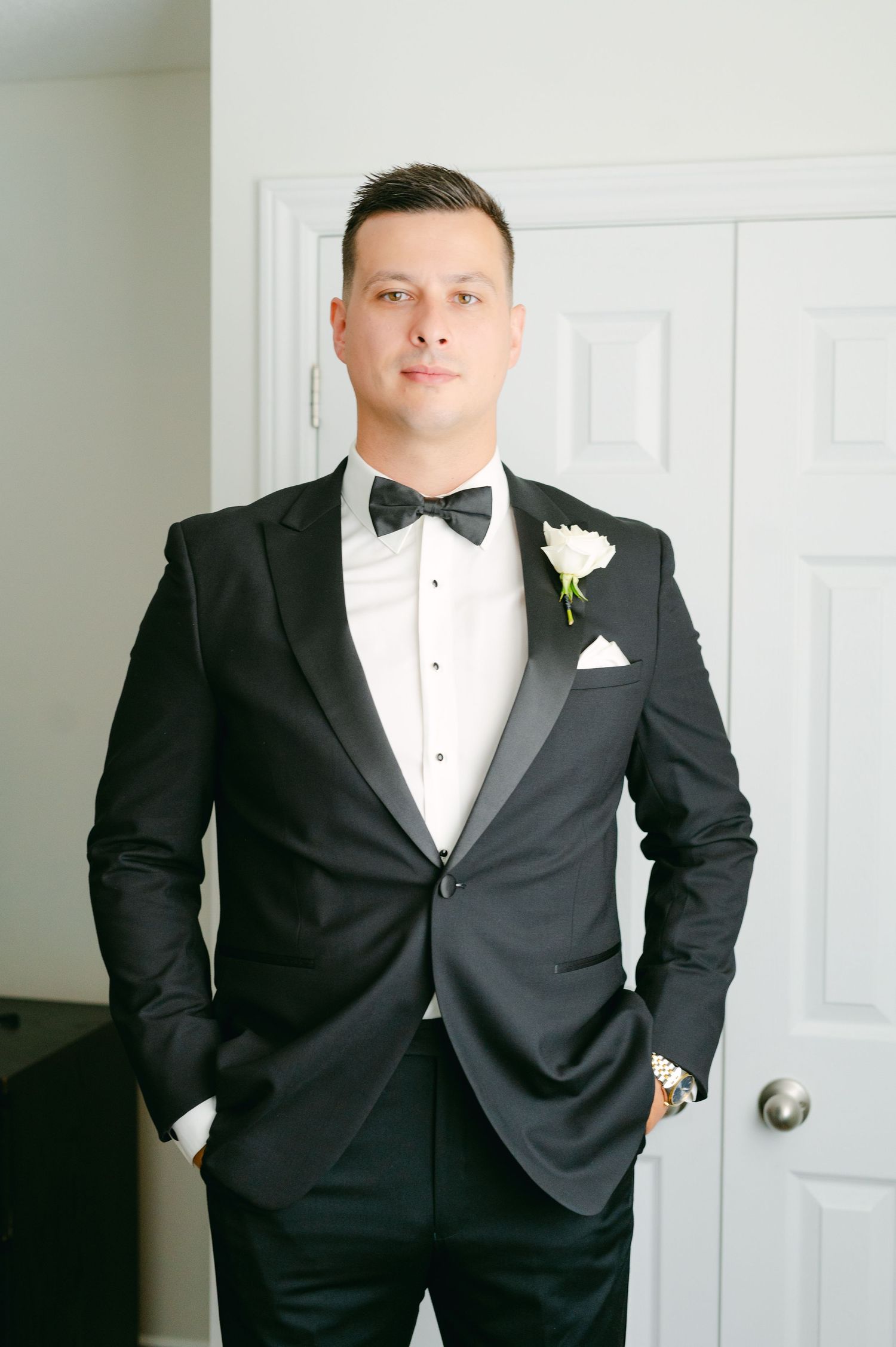 A formal portrait of a groom in a black tuxedo with white boutonniere against a white door.
