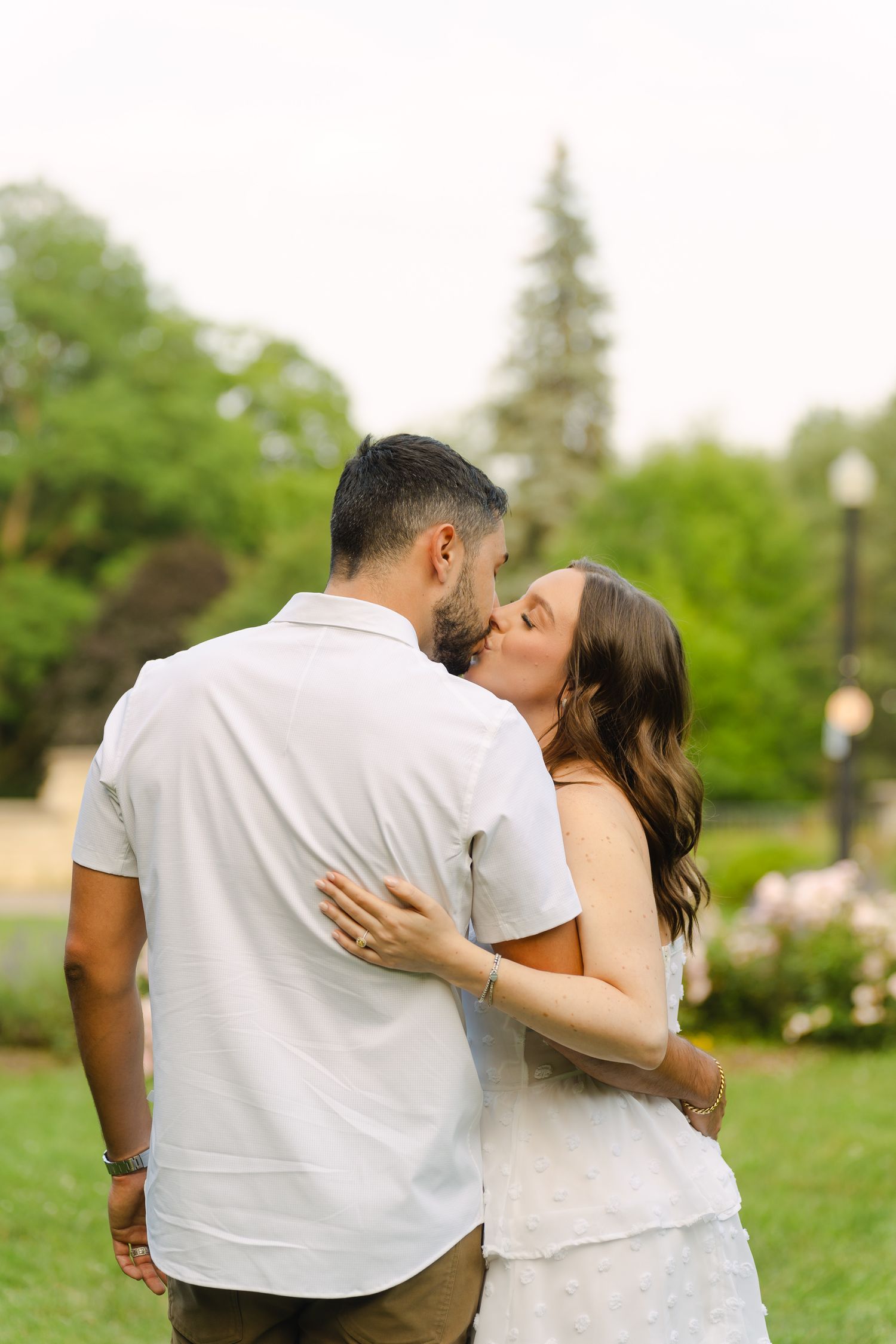 A loving couple embraces in a garden setting surrounded by green trees and blooming flowers.