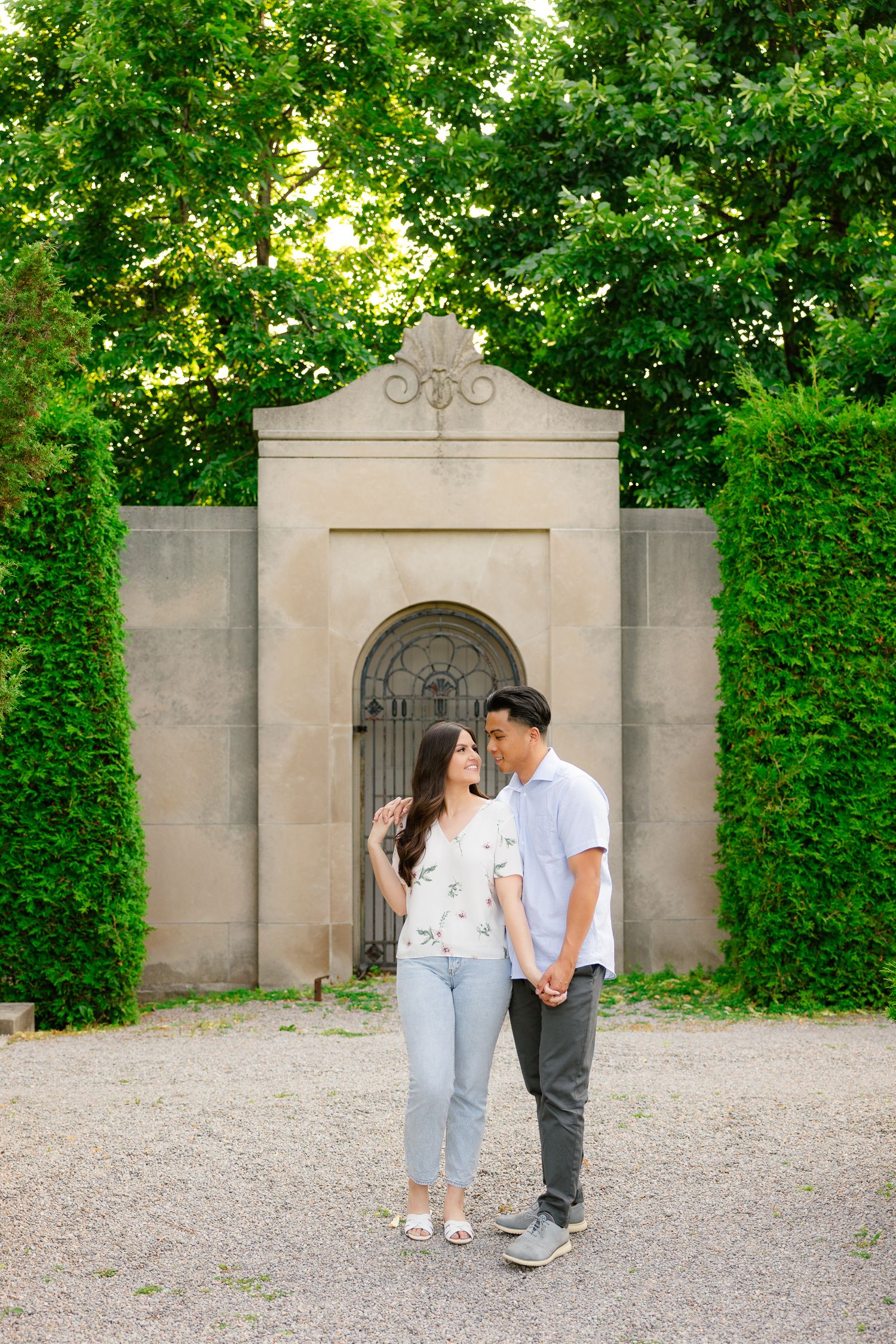 A couple poses in front of an ornate stone archway surrounded by manicured hedges.