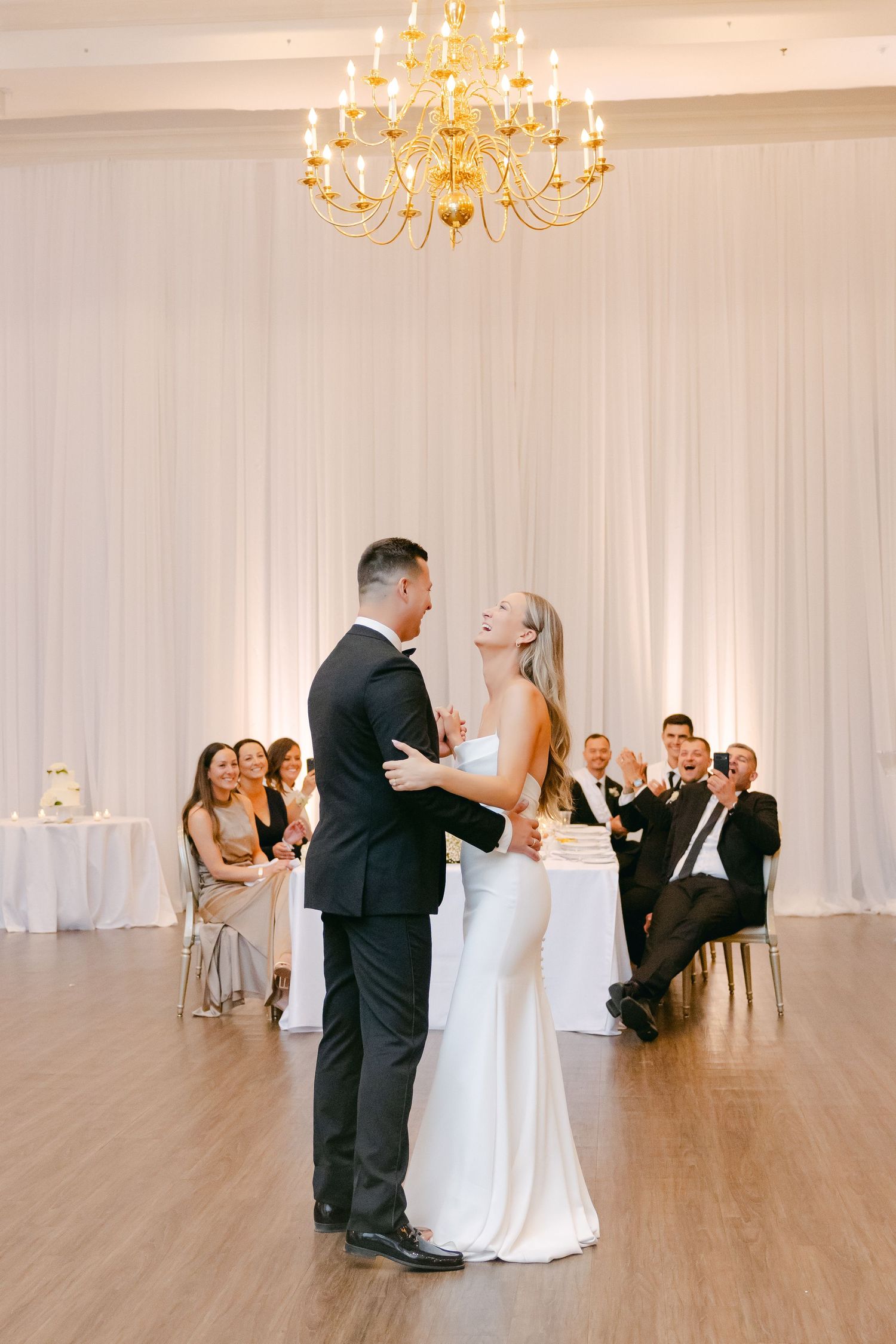 A couple shares their first dance at a wedding reception under an elegant gold chandelier in a white-draped ballroom.