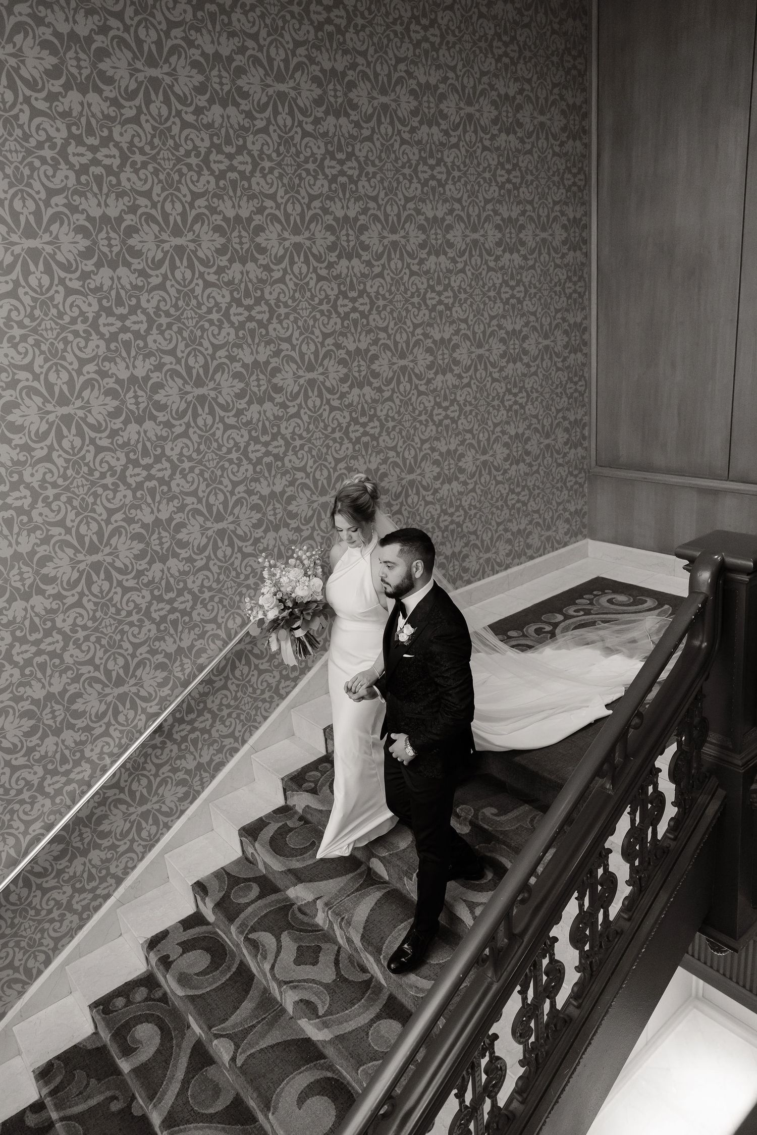 Wedding couple descending a grand staircase with patterned wallpaper in black and white.