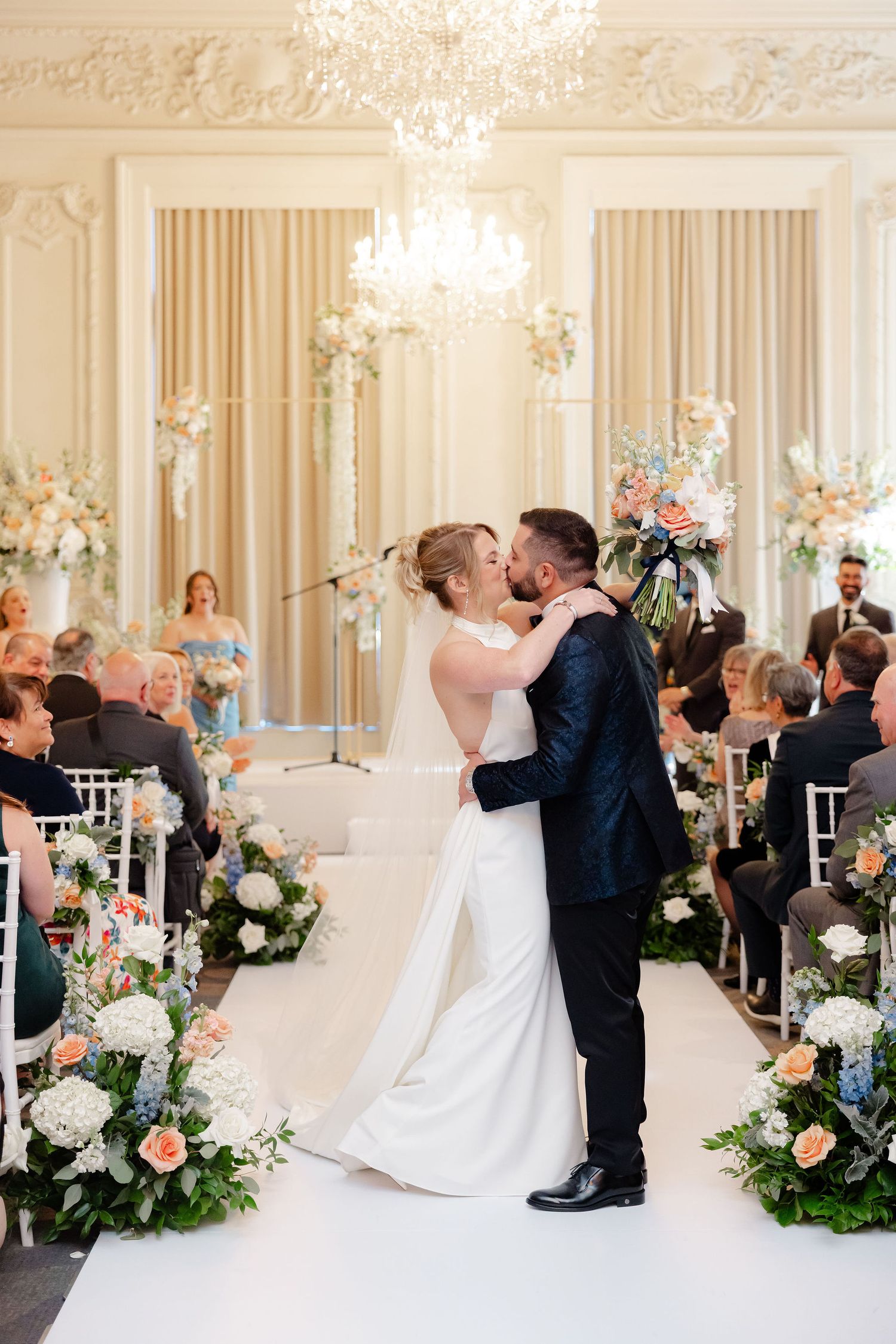 Wedding ceremony moment in an ornate white ballroom with floral arrangements and white chairs.