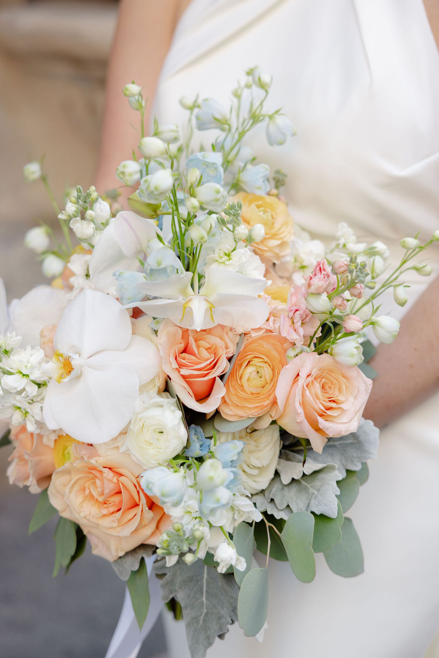 A romantic wedding bouquet with peach roses, white orchids, and delicate blue flowers mixed with eucalyptus foliage.