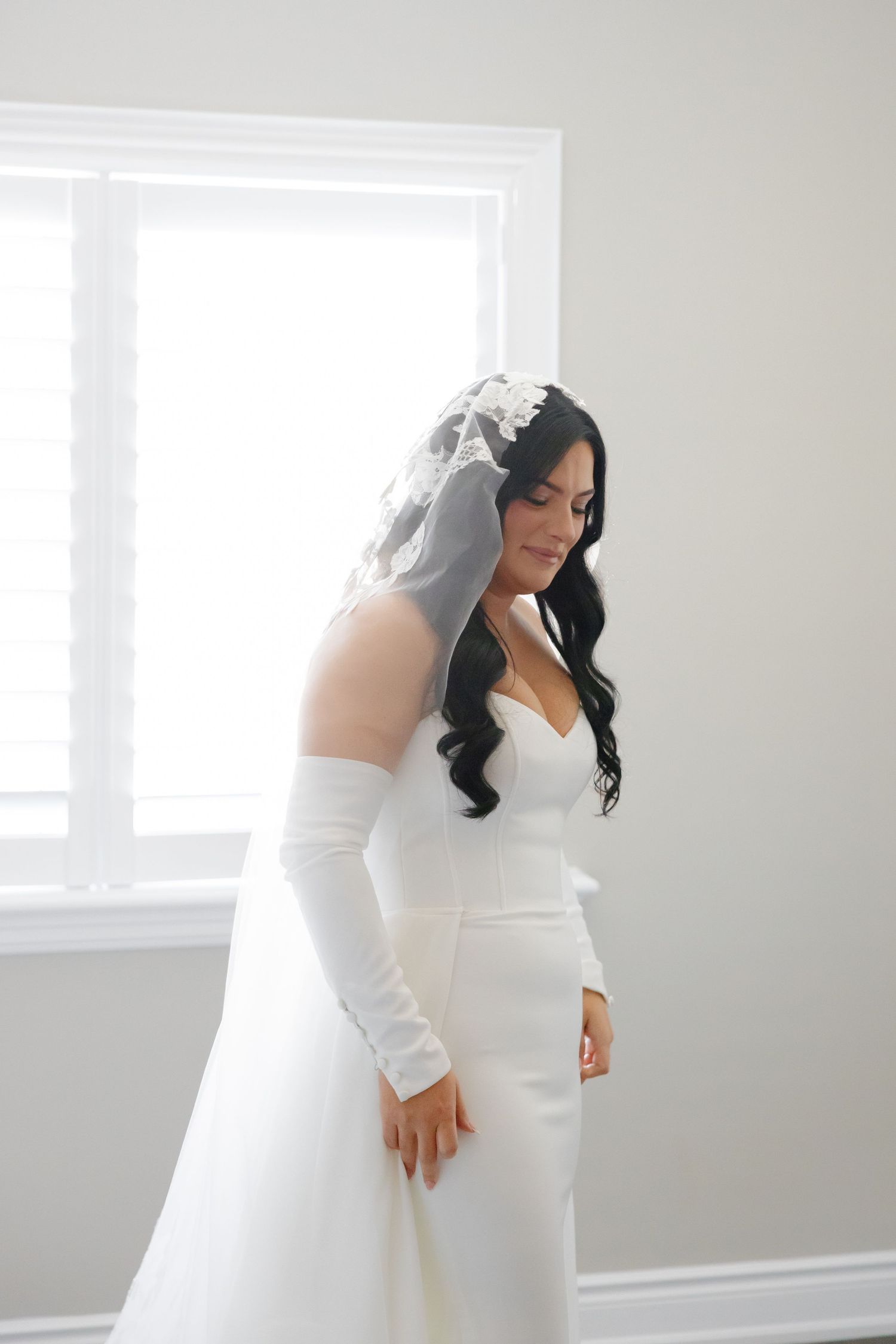 A bride in an elegant fitted white gown and veil stands in profile by a bright window.