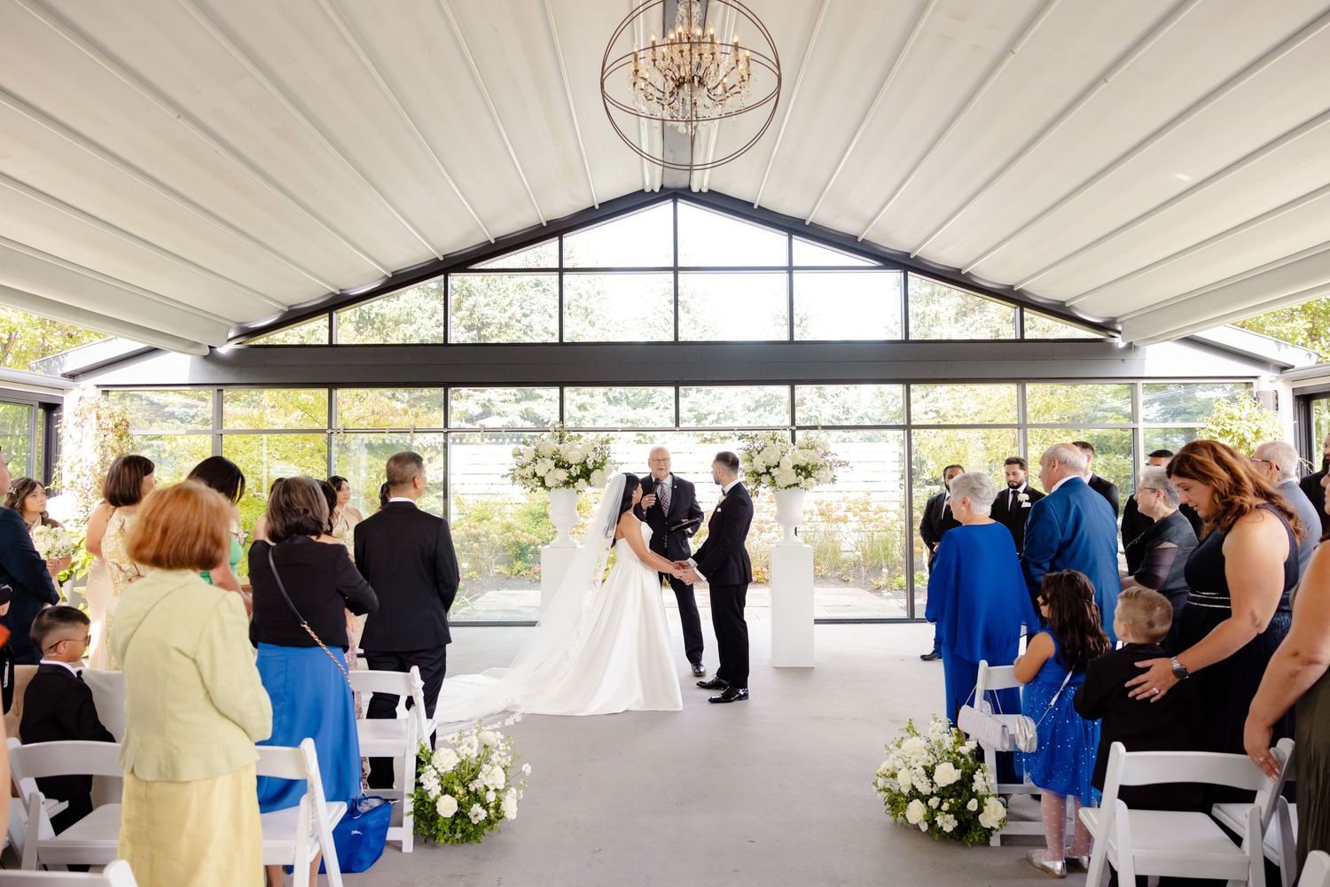 Wedding ceremony in sunlit venue with guests seated in white chairs and blue attire.