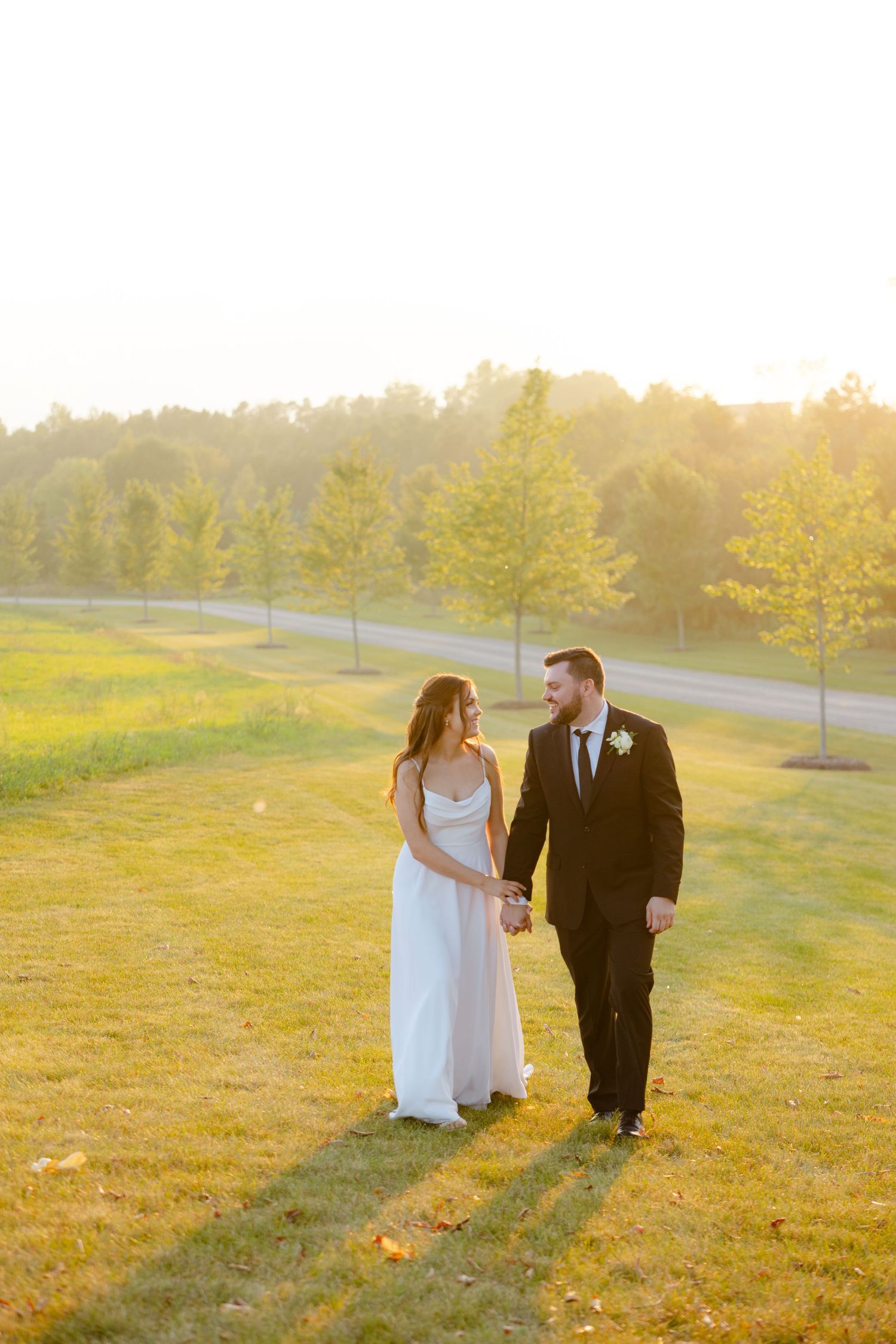 A couple shares a loving glance while walking hand in hand on a sunlit grassy hill.