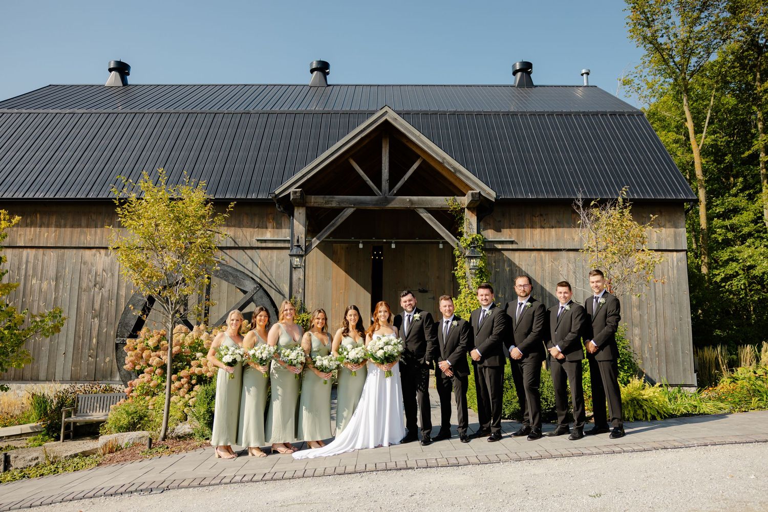 Wedding party poses in front of a rustic barn venue with sage green bridesmaid dresses and black suits.