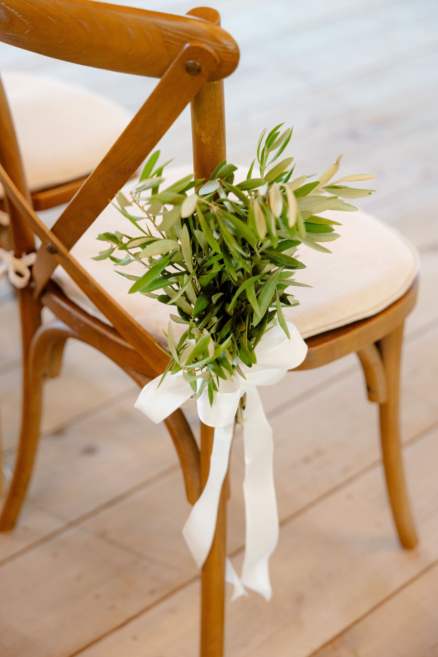 Wooden cross-back chair decorated with delicate greenery and white ribbon for wedding ceremony.
